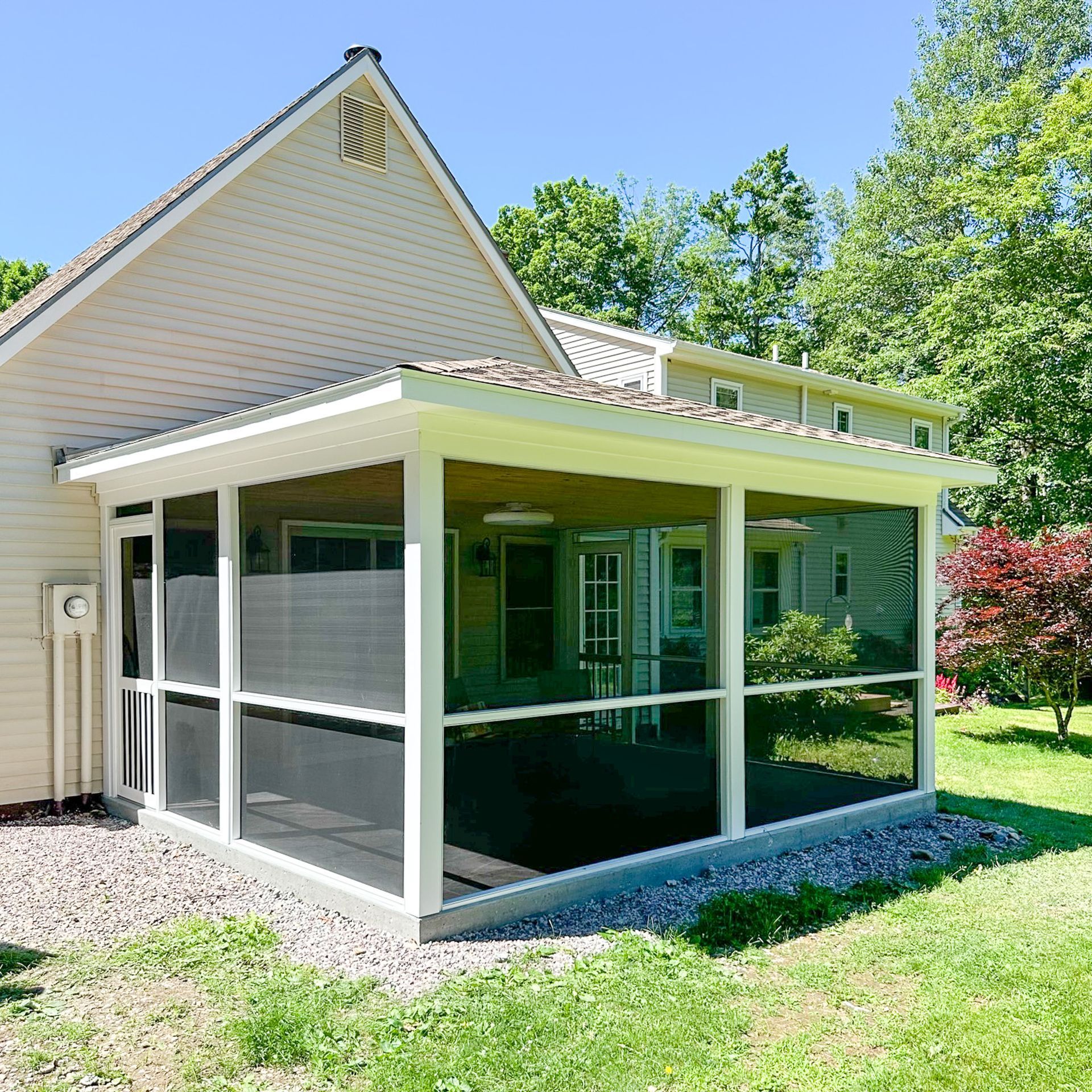 Screened-in porch attached to a light-colored house, with white trim and black screening. A grassy yard and trees surround it.