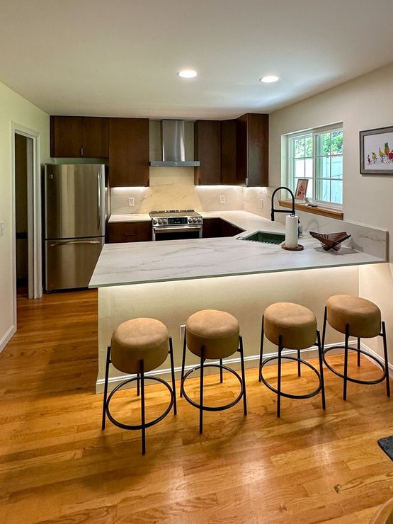 Modern kitchen with brown cabinets, a white countertop, stainless steel appliances, and four stools at the counter.