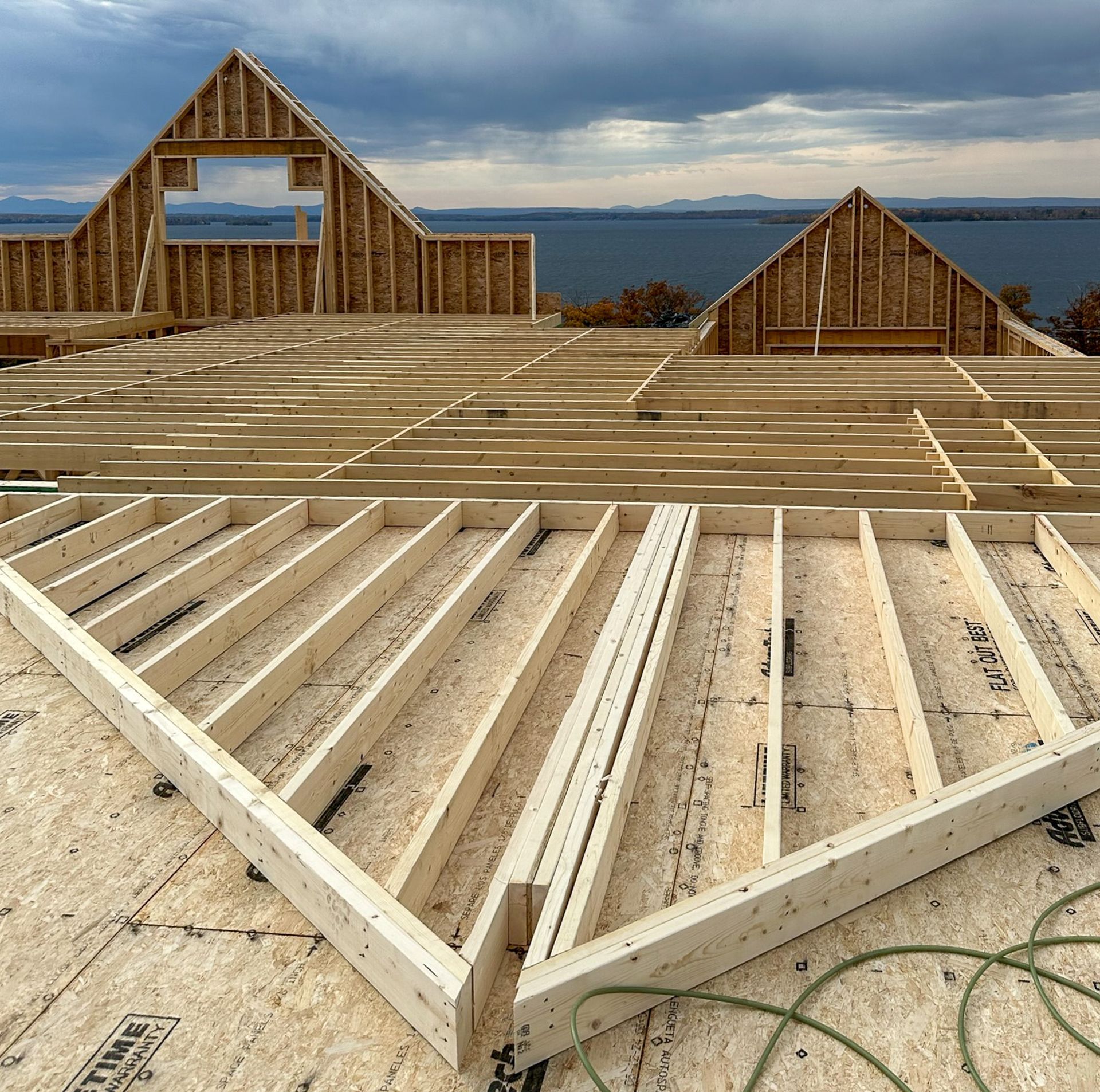 Wooden house under construction with exposed framing, against a cloudy sky and a body of water.