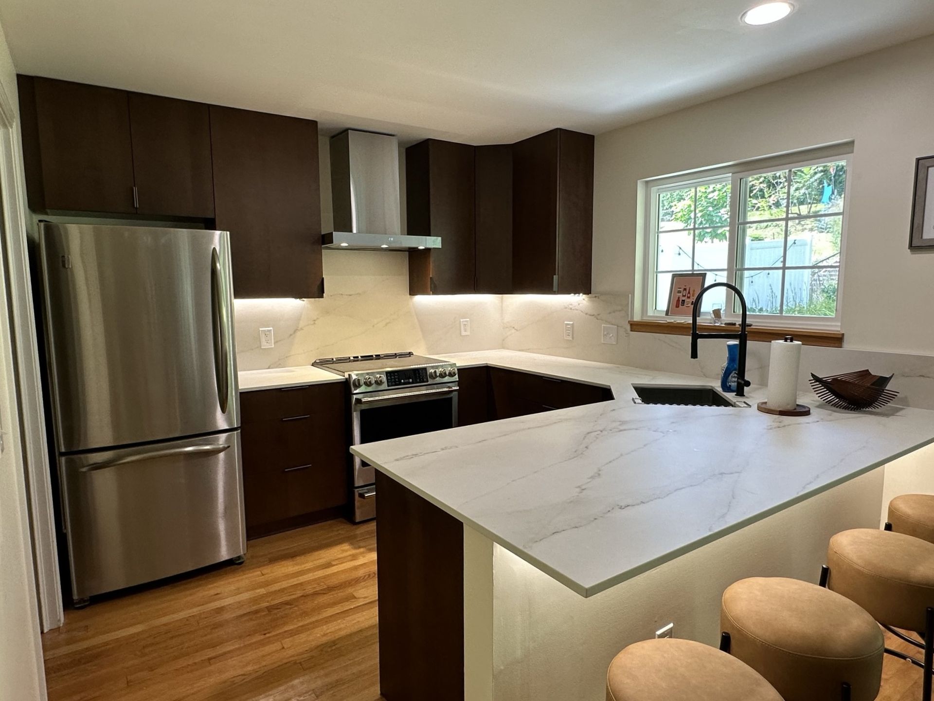 Modern kitchen with dark brown cabinets, stainless steel appliances, white countertops, and a breakfast bar with tan stools.
