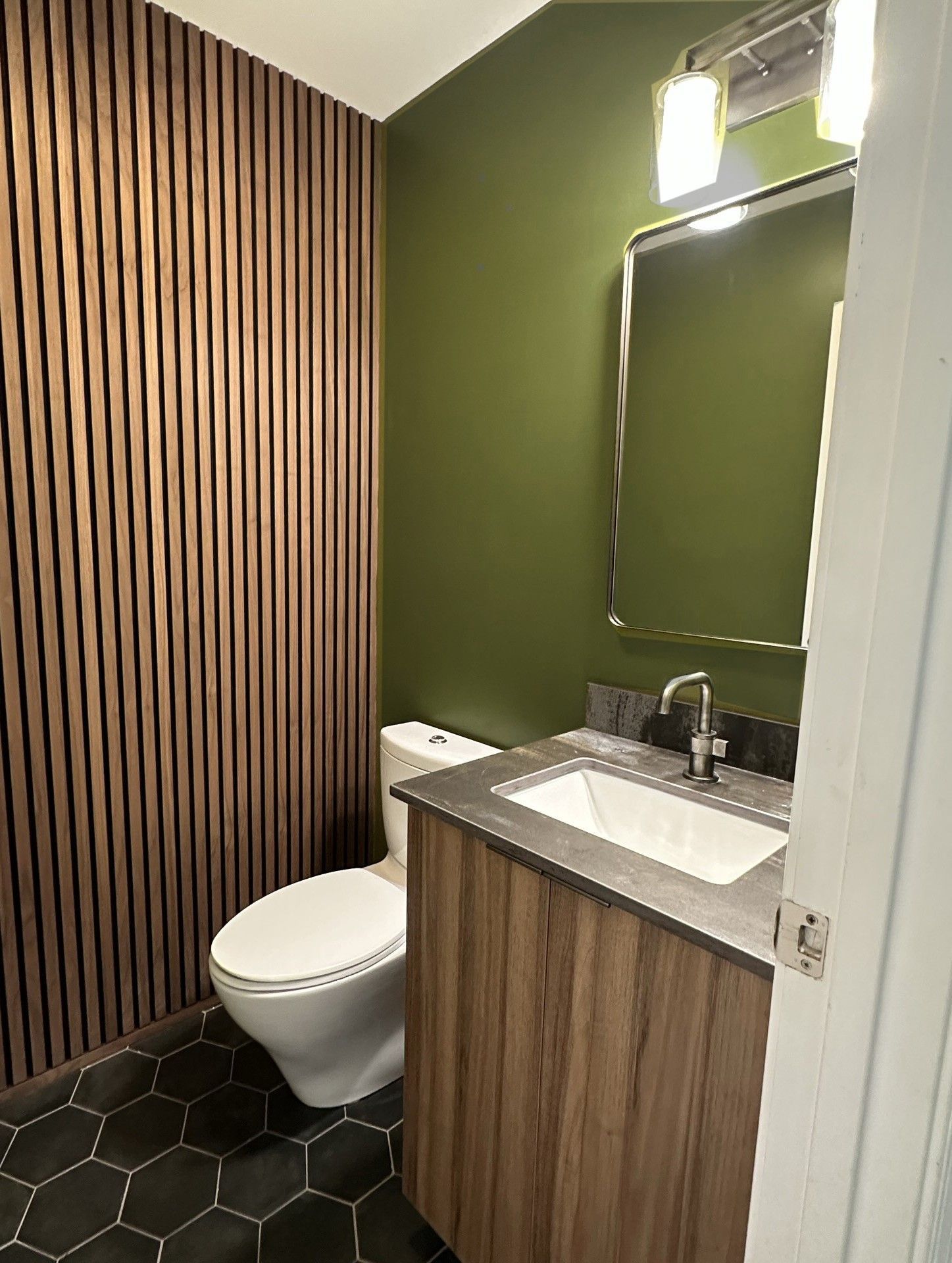 Small bathroom with a wooden slatted wall, green wall, gray countertop, dark hexagonal tile floor.