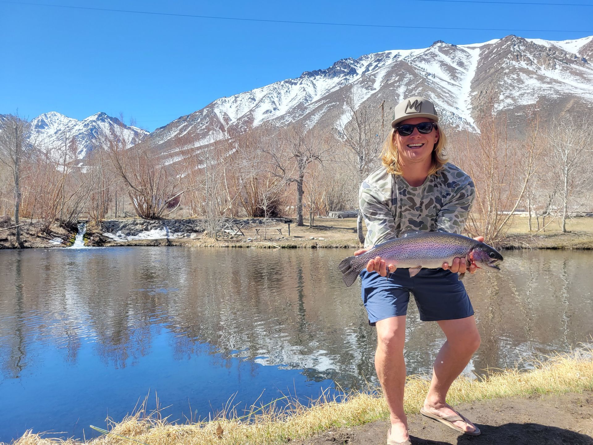 A woman is holding a fish in front of a lake with mountains in the background.