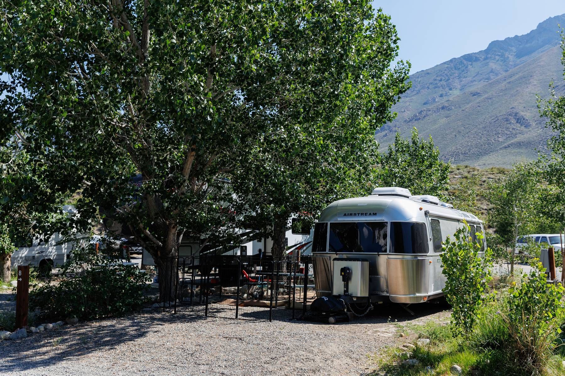 A silver airstream trailer is parked in a gravel lot with trees and mountains in the background.