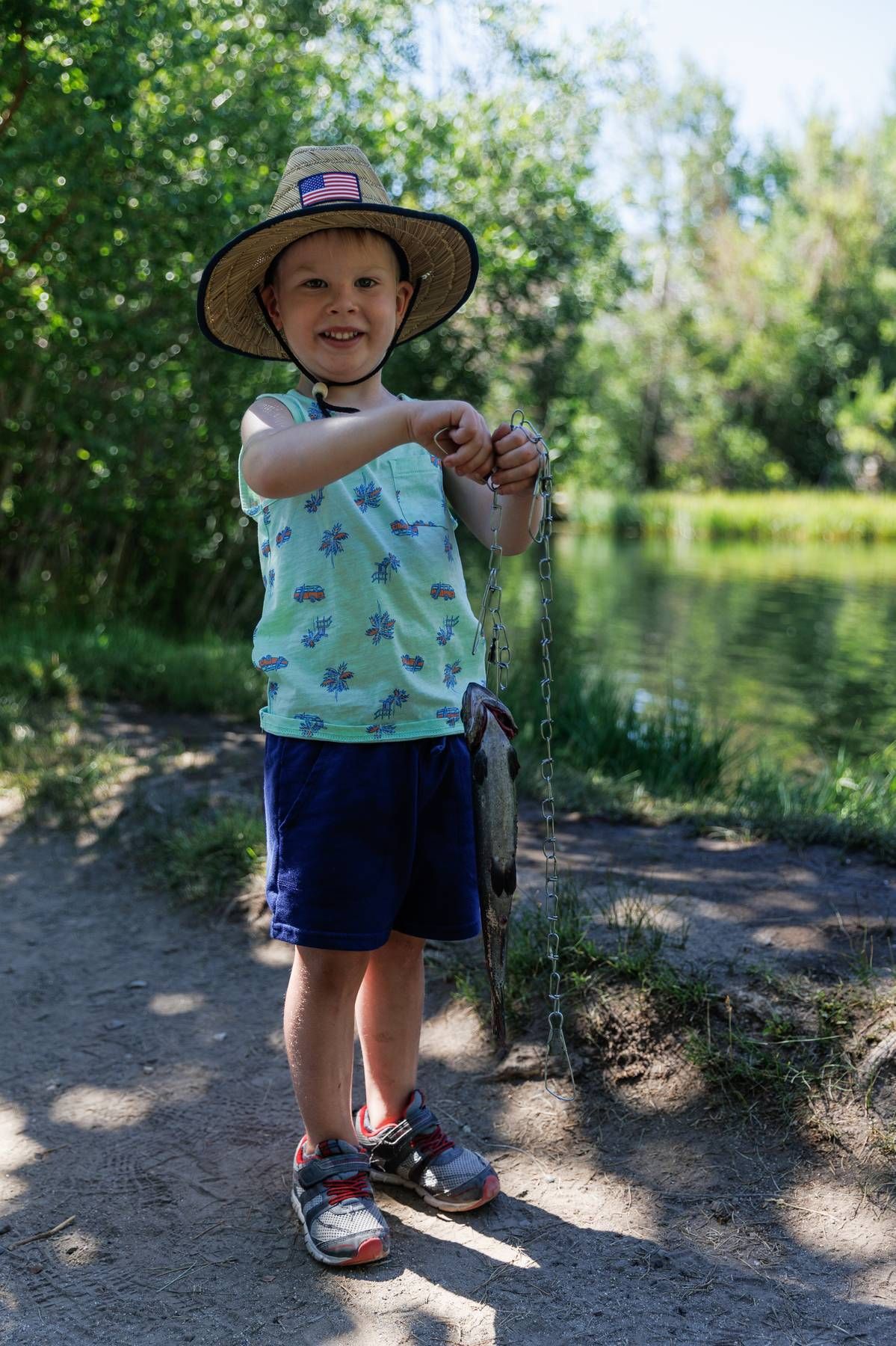 A young boy is standing next to a lake holding a fish on a fishing rod.