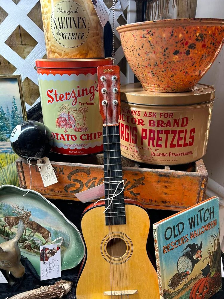 Vintage kitchen decor with stacked tins, a metal colander, pretzel can, guitar, and an “Old Witch” tin sign
