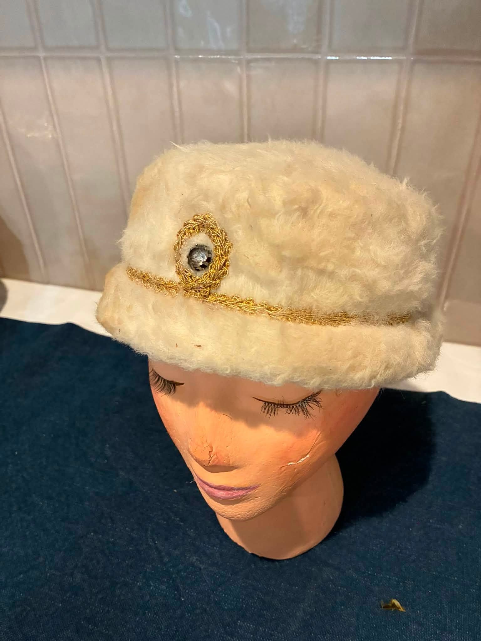 Fuzzy cream-colored hat with gold trim and a jeweled emblem on a mannequin head.