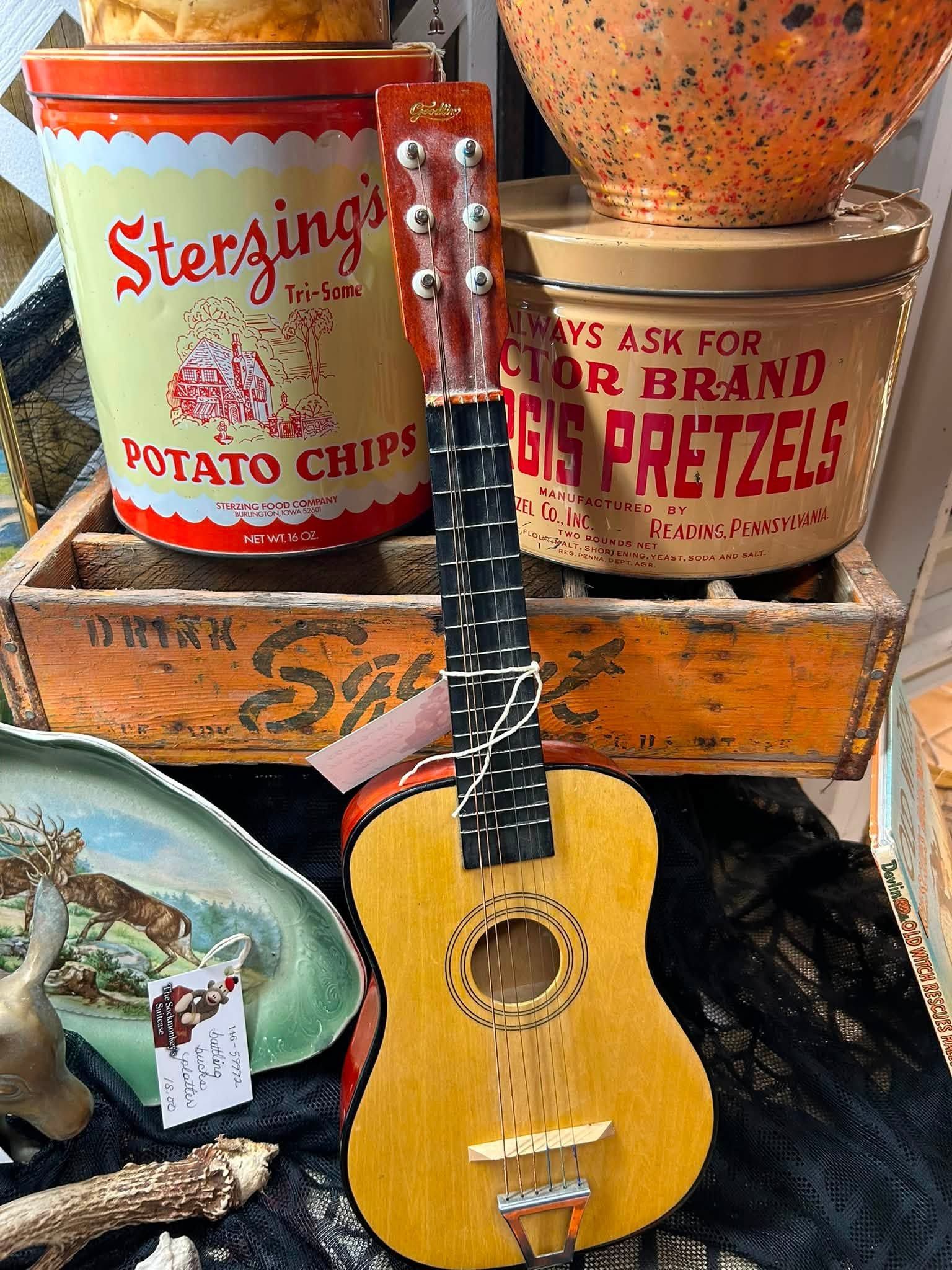 Small guitar on stacked vintage snack tins and a crate, with pretzels and potato chips in the background