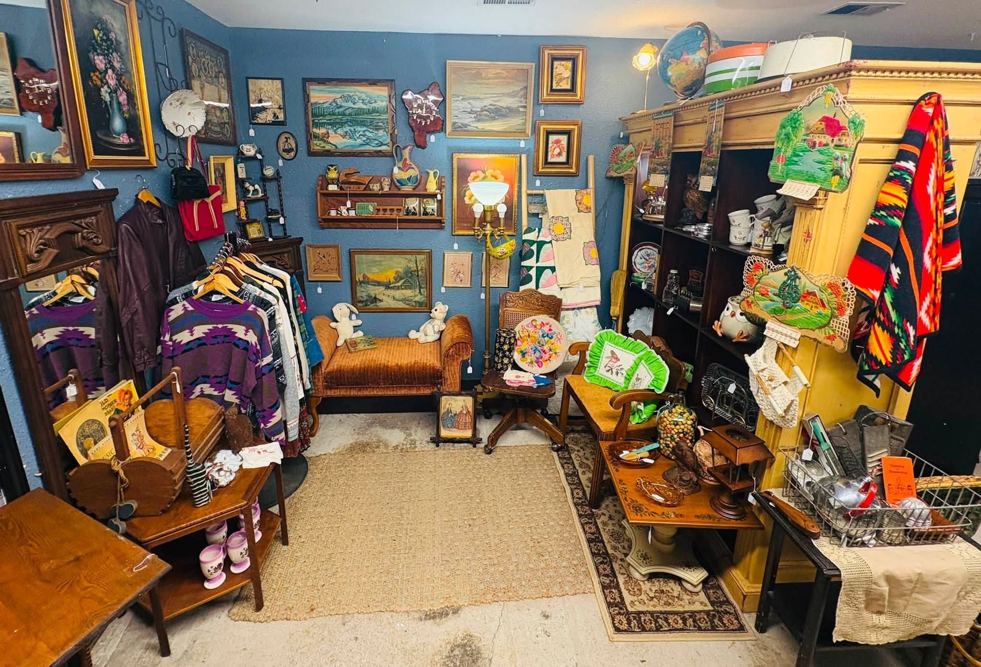 Cluttered thrift shop or vintage boutique with colorful clothes, paintings, and household items on display.