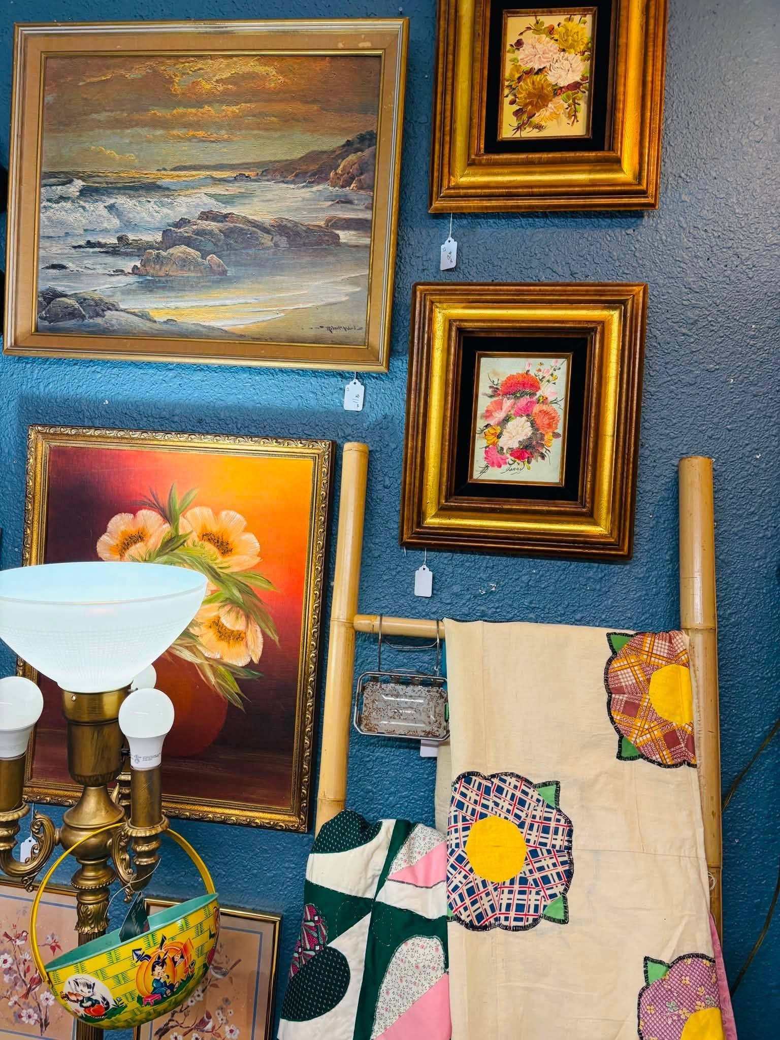 Assorted framed paintings and quilts displayed on a blue wall, with a floor lamp in the foreground.