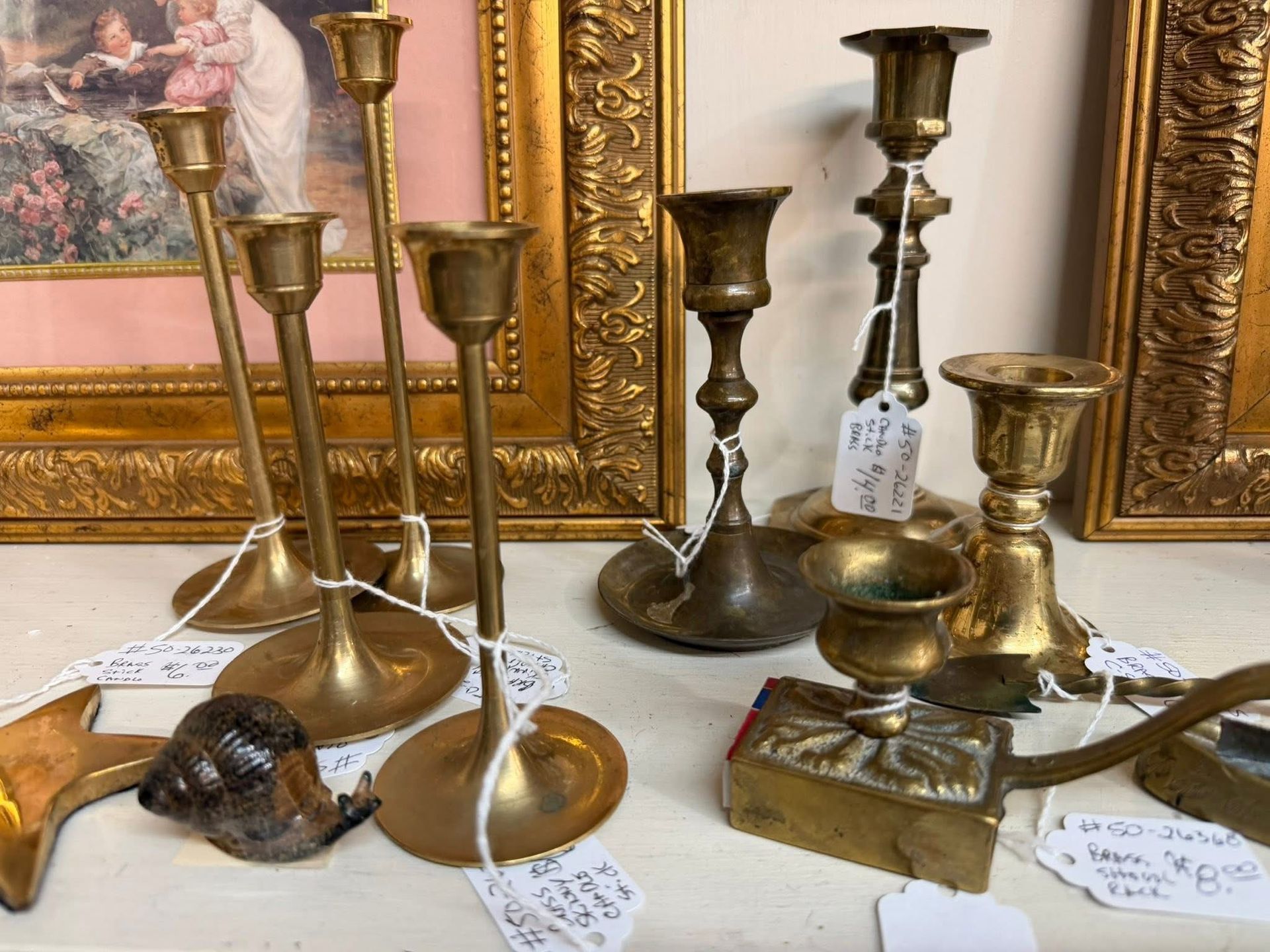Assorted brass and bronze candlesticks and decorative items arranged on a table in front of framed paintings