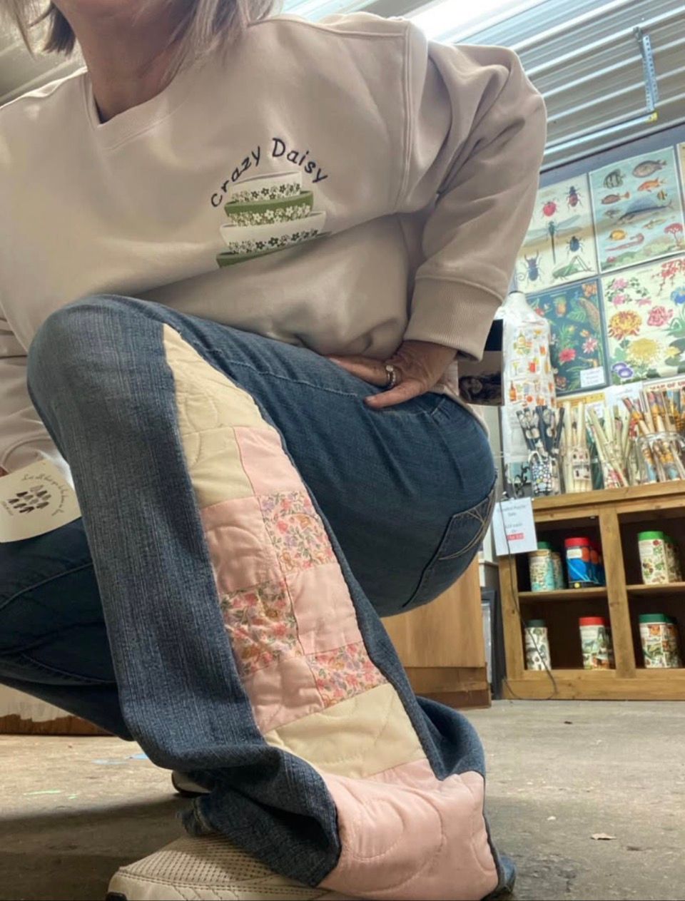 Person sitting indoors in a beige sweatshirt and blue jeans with pink patchwork legs in a store.