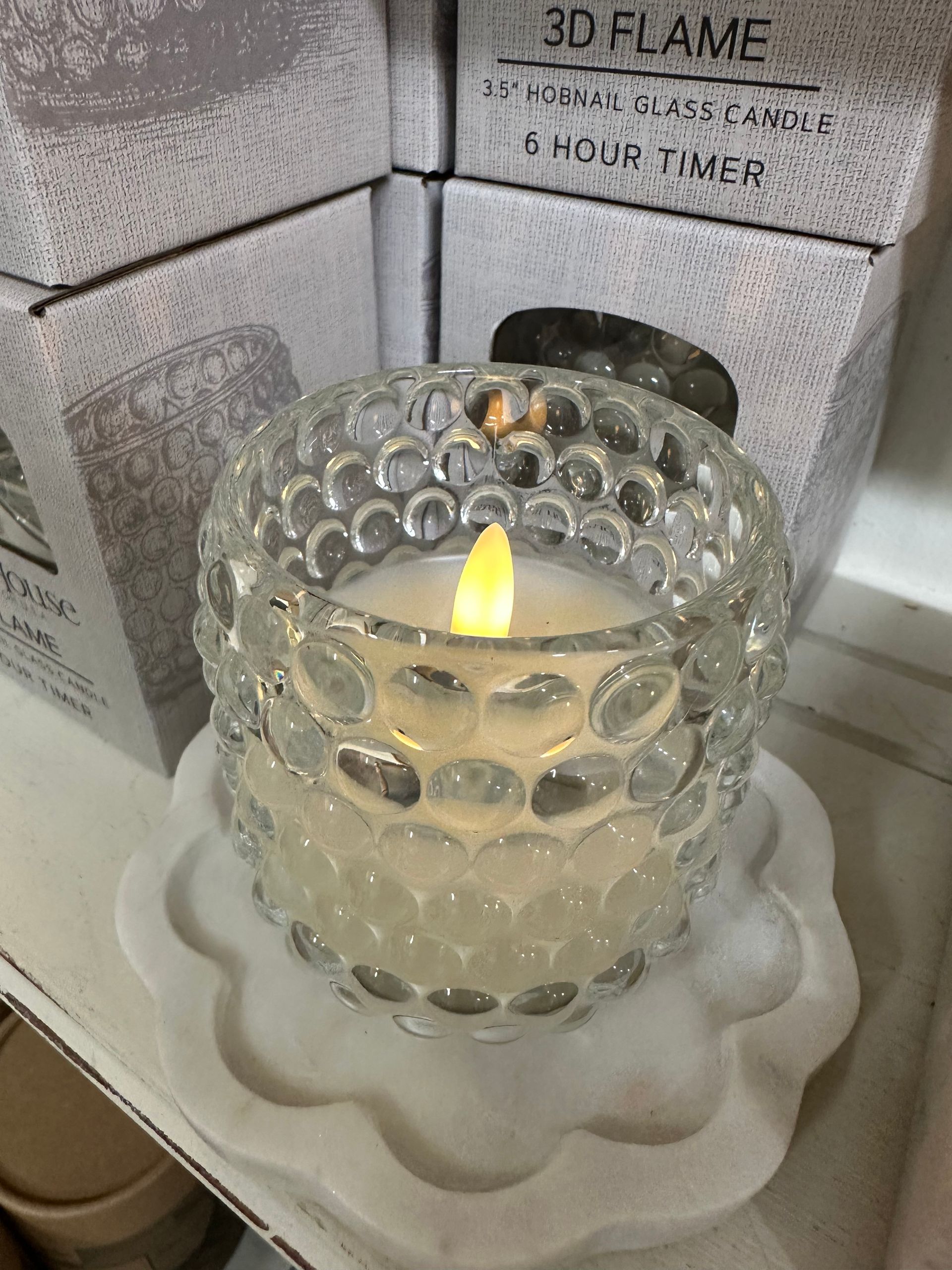 Lit candle in a clear textured glass holder on a white saucer, with boxes in the background