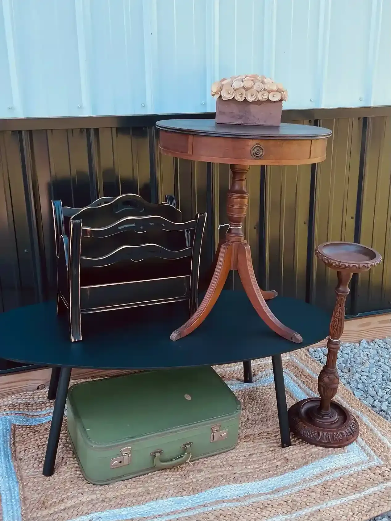 Miniature patio display with wooden table, chair, small rug, green suitcase, and flower pot on a shelf