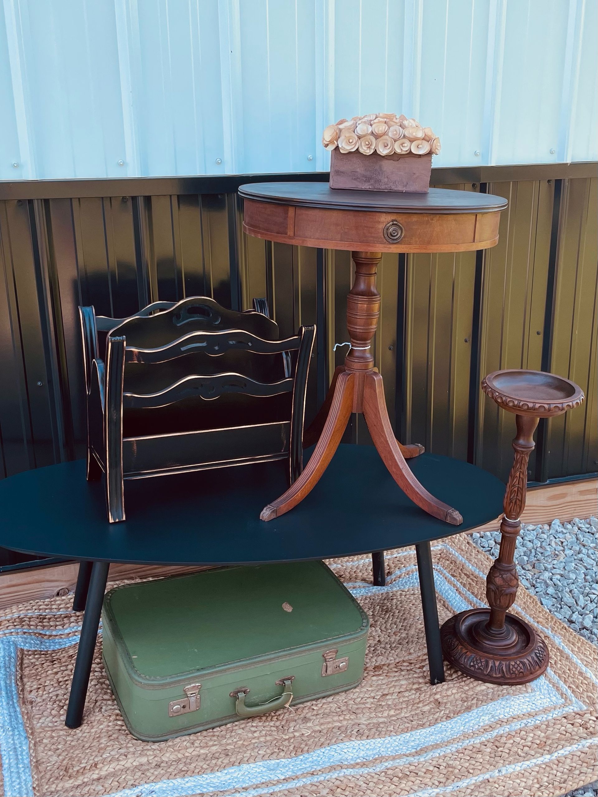 Miniature patio display with wooden table, chair, small rug, green suitcase, and flower pot on a shelf