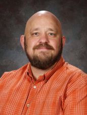 A bald man with a beard is wearing an orange plaid shirt and smiling for the camera.