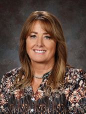 A woman wearing glasses and overalls is smiling for a picture.