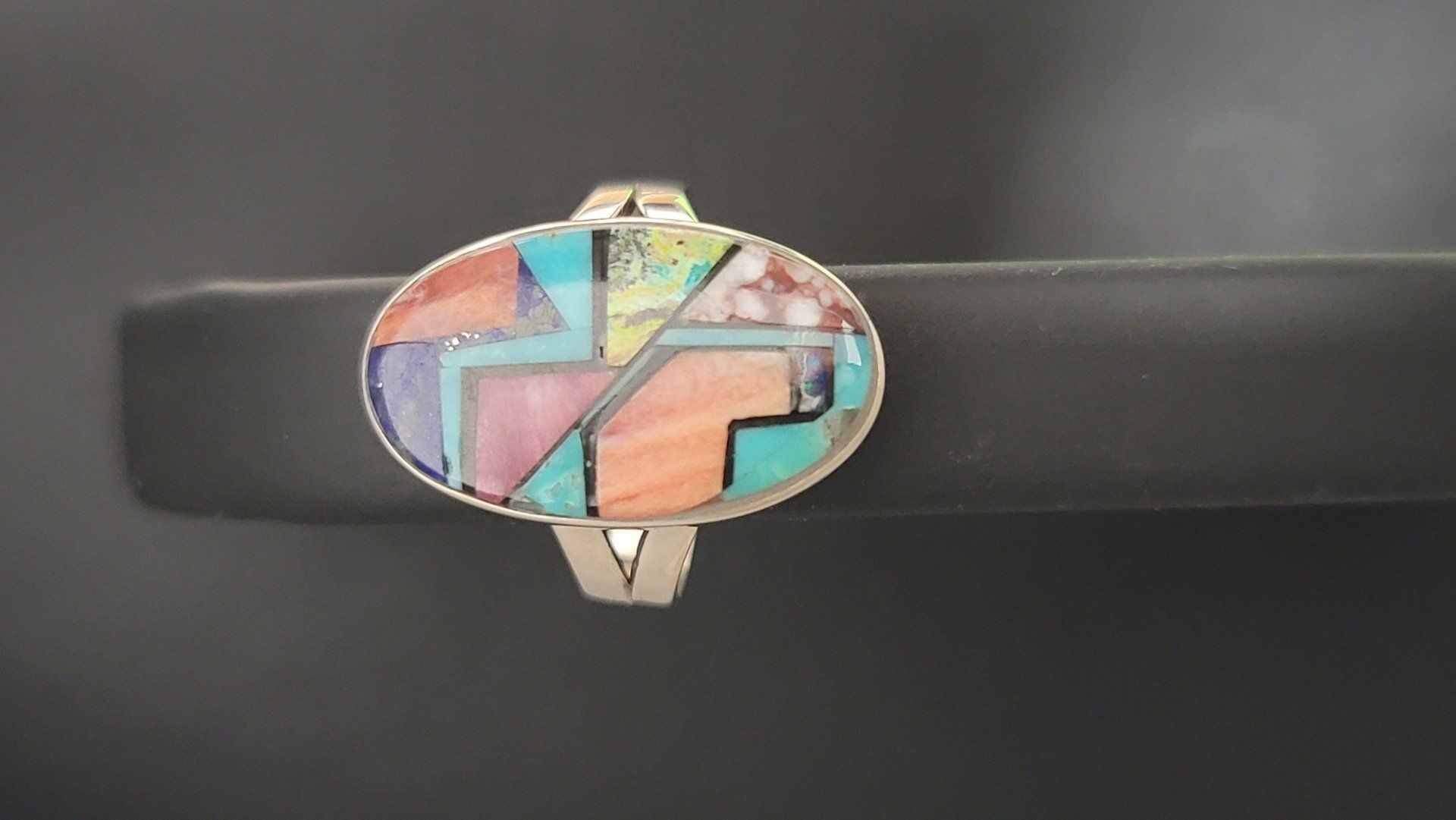 Oval-shaped silver ring with inlaid colorful stones arranged in geometric patterns.