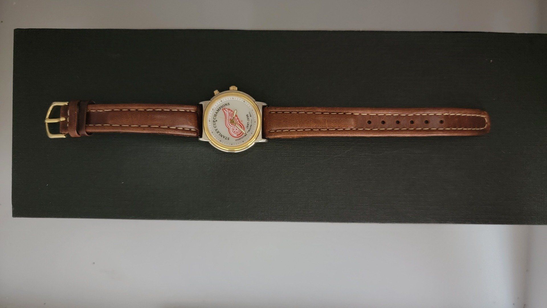 A brown leather watch with a gold-colored case and face displaying red and white markings, on a dark surface.