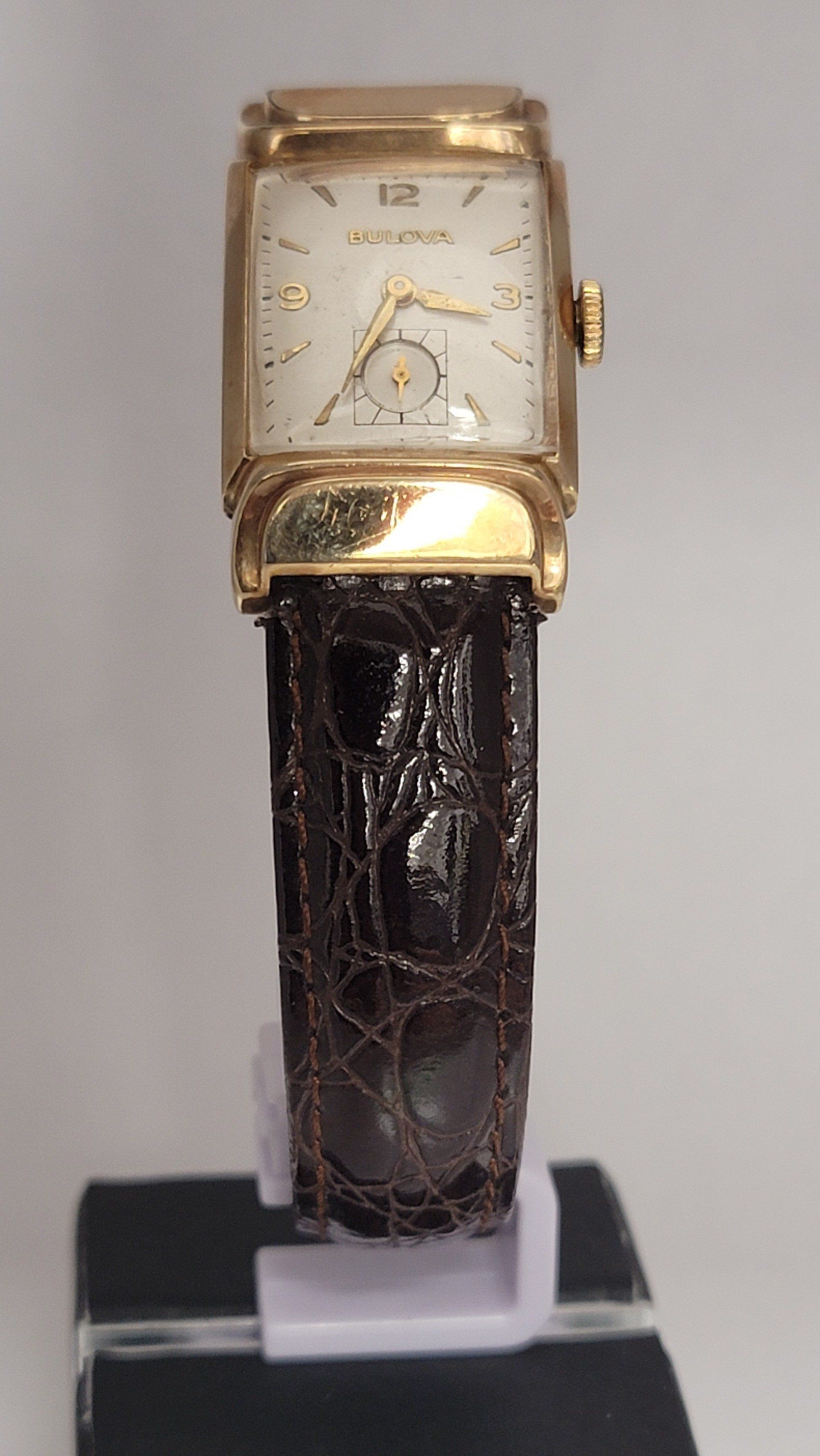 Gold rectangular wristwatch with a brown leather strap displayed on a stand.