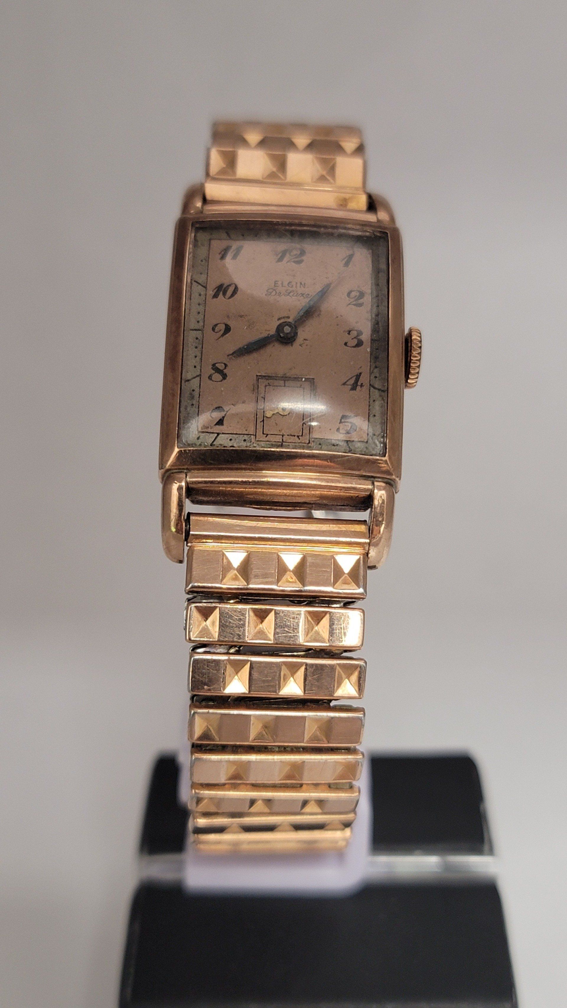 Gold-toned rectangular wristwatch with a patterned band and face, sitting on a black stand.