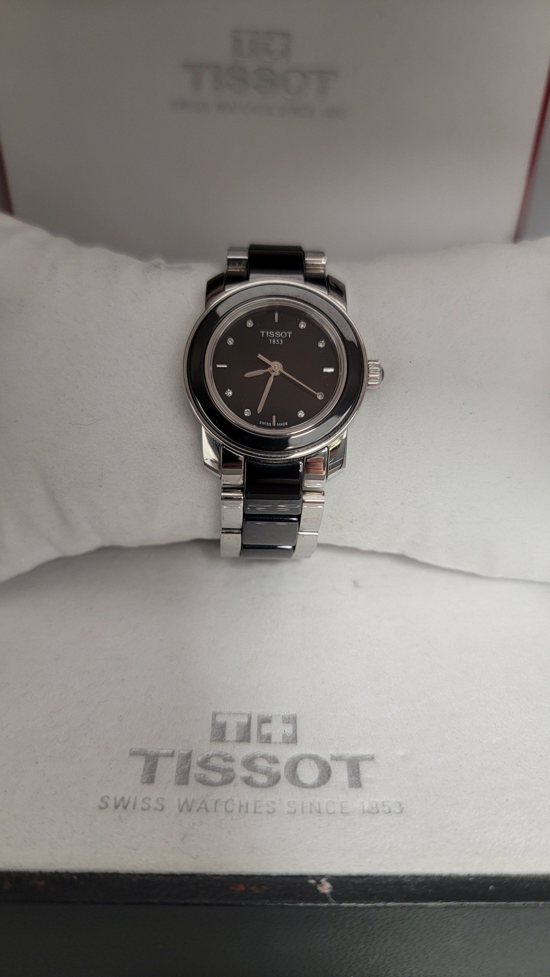 Tissot watch with black face and bezel, silver and black band, in its branded box on a white pillow.
