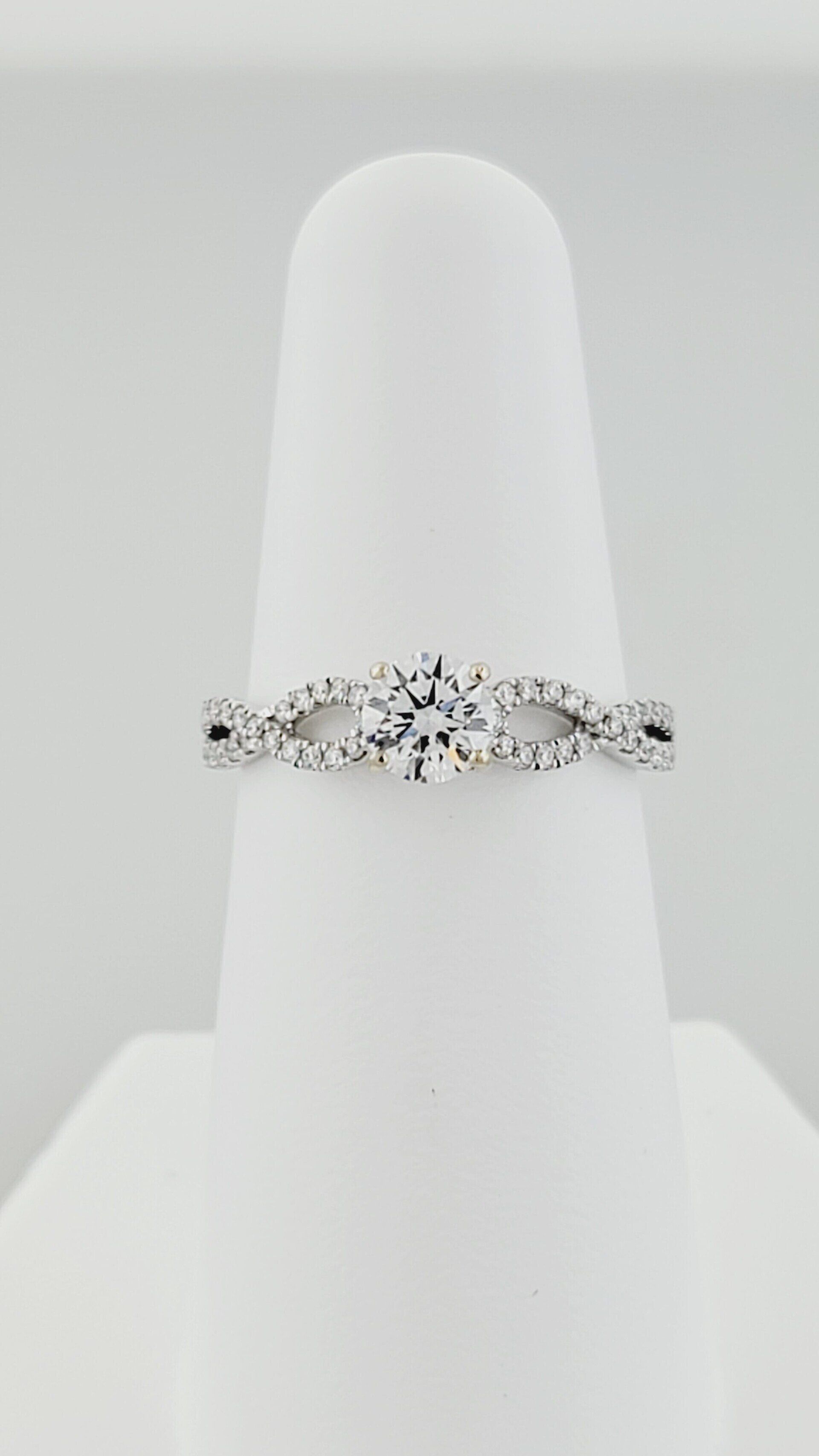 Diamond engagement ring, featuring a round center stone and a twisted band with smaller diamonds, on a white display stand.