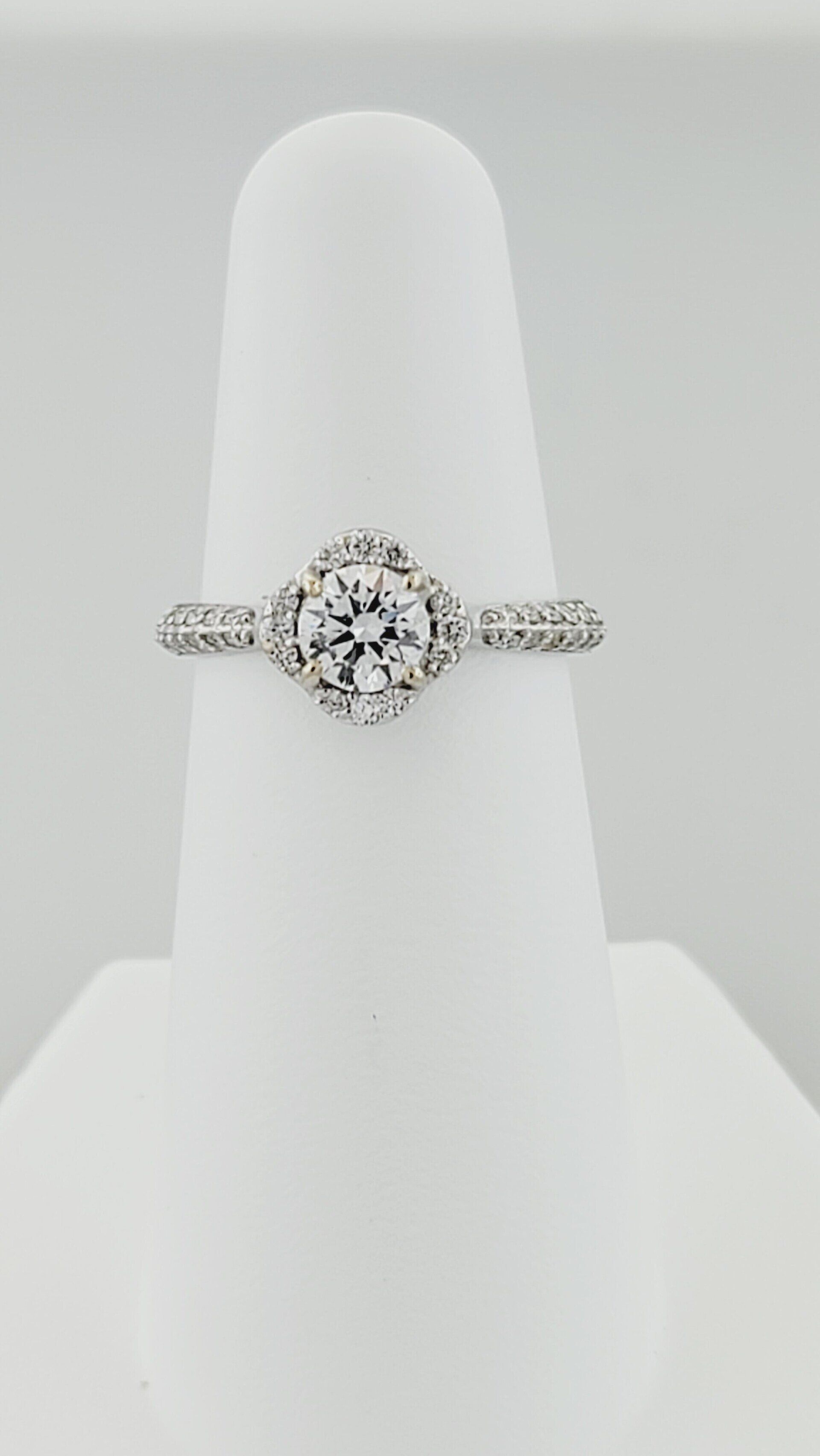 Diamond engagement ring on white finger-shaped display, with halo setting and pave band, against white background.