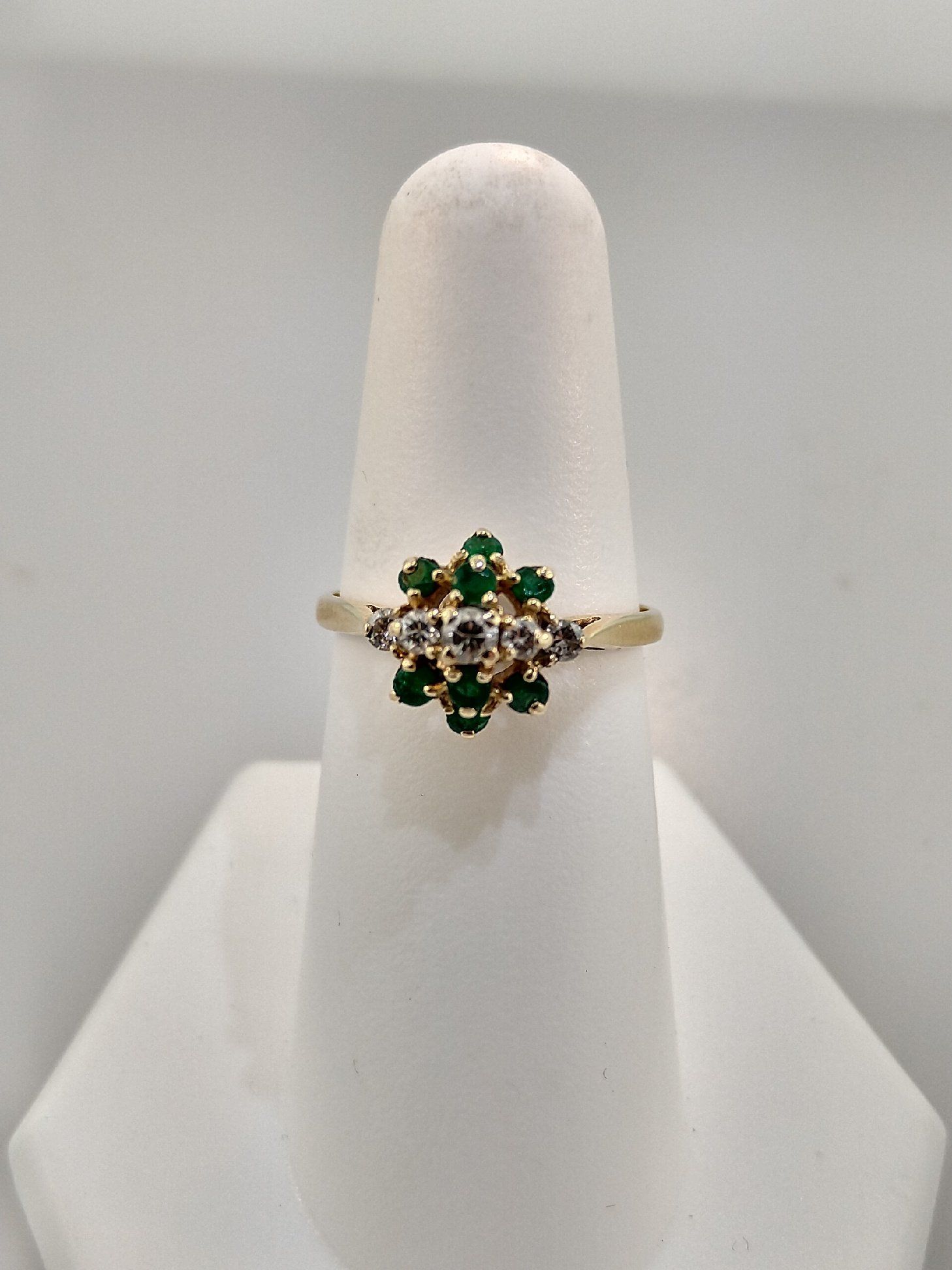 Gold ring with a flower-shaped setting of emeralds and diamonds on a white display stand.