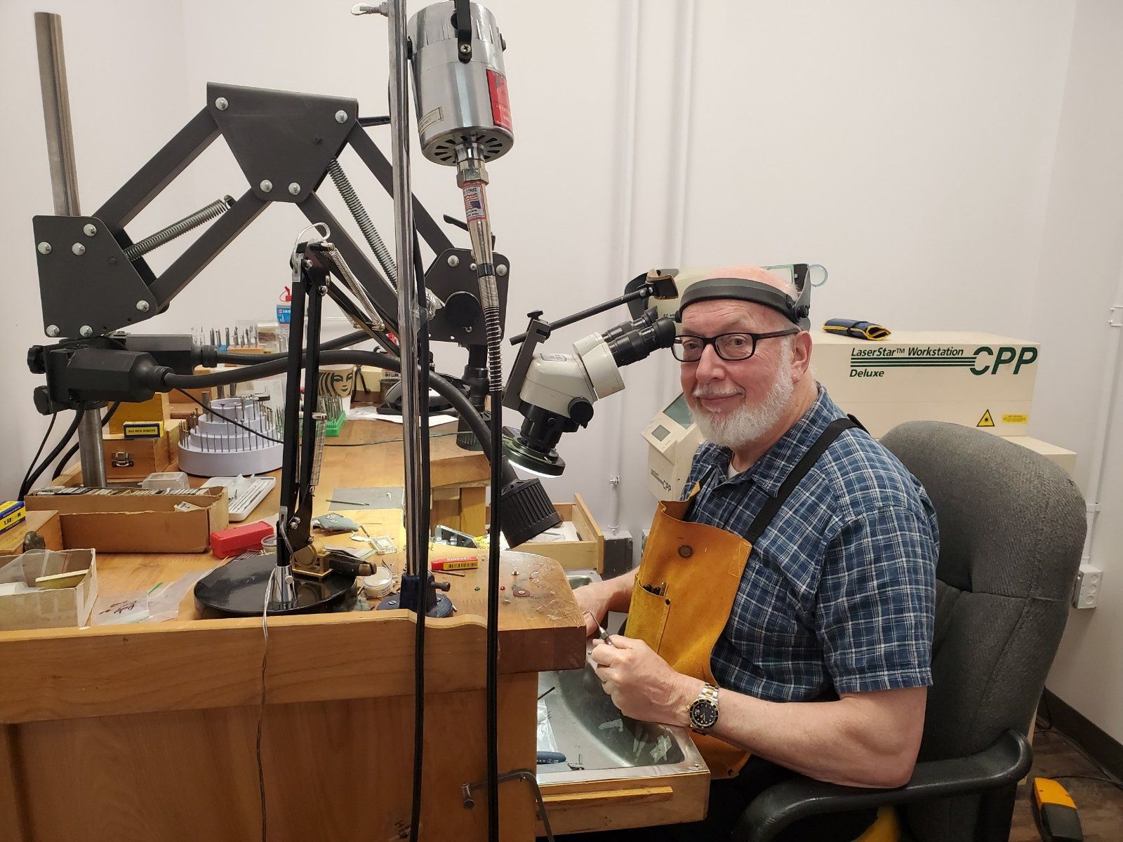 Man working at a workbench with jewelry-making tools, wearing safety glasses and apron.