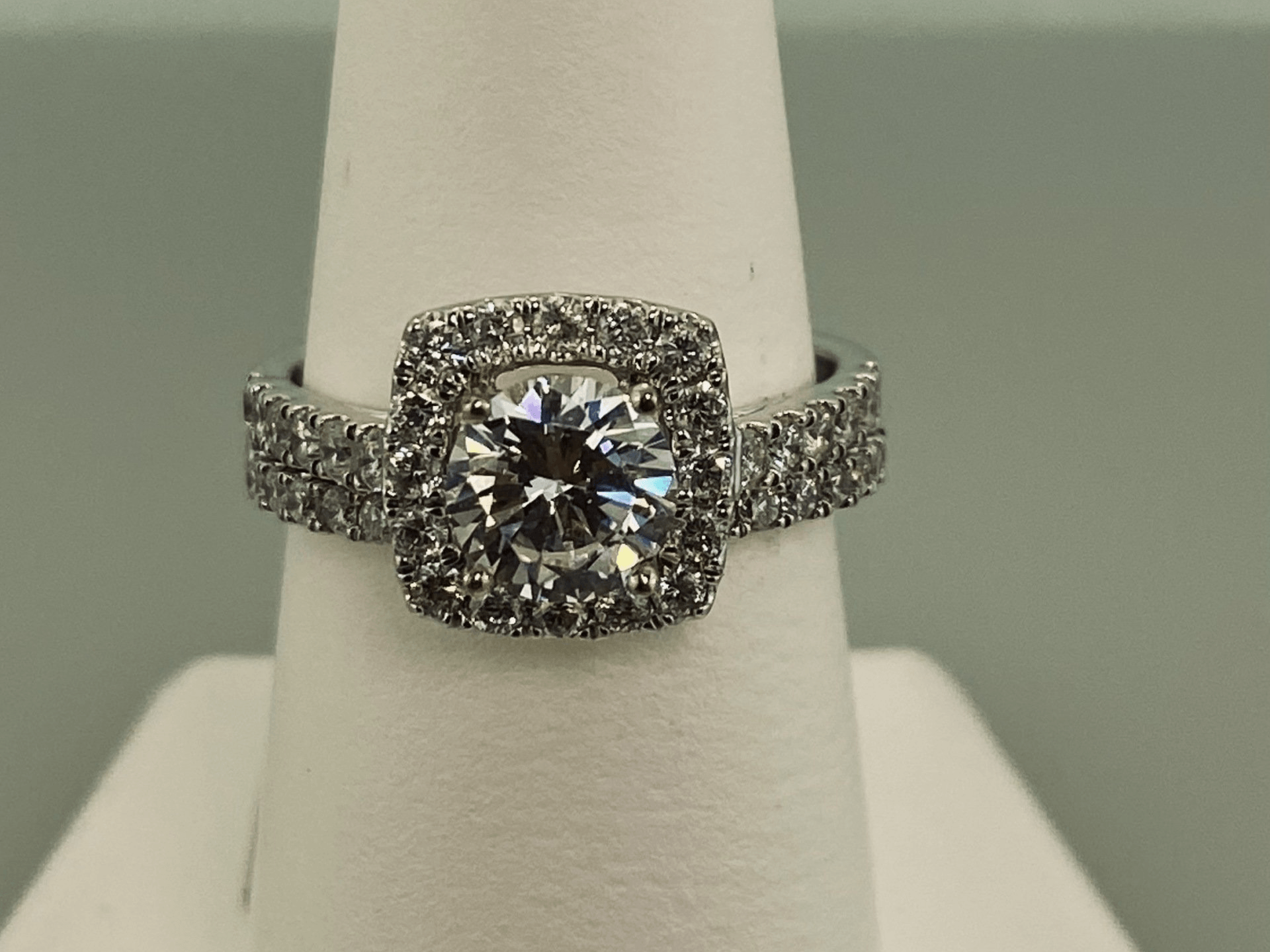 Diamond ring with a square halo and band, on a white display stand.