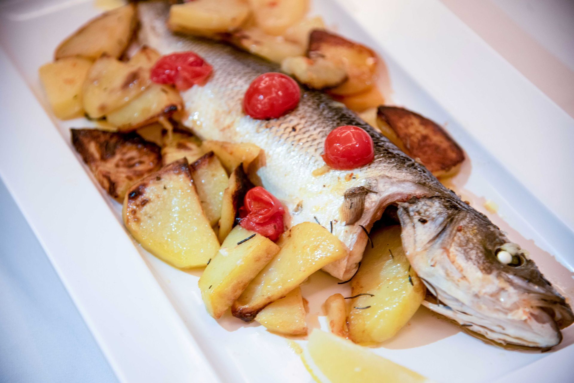 Pesce intero al forno con patate arrosto e pomodorini su un piatto bianco.