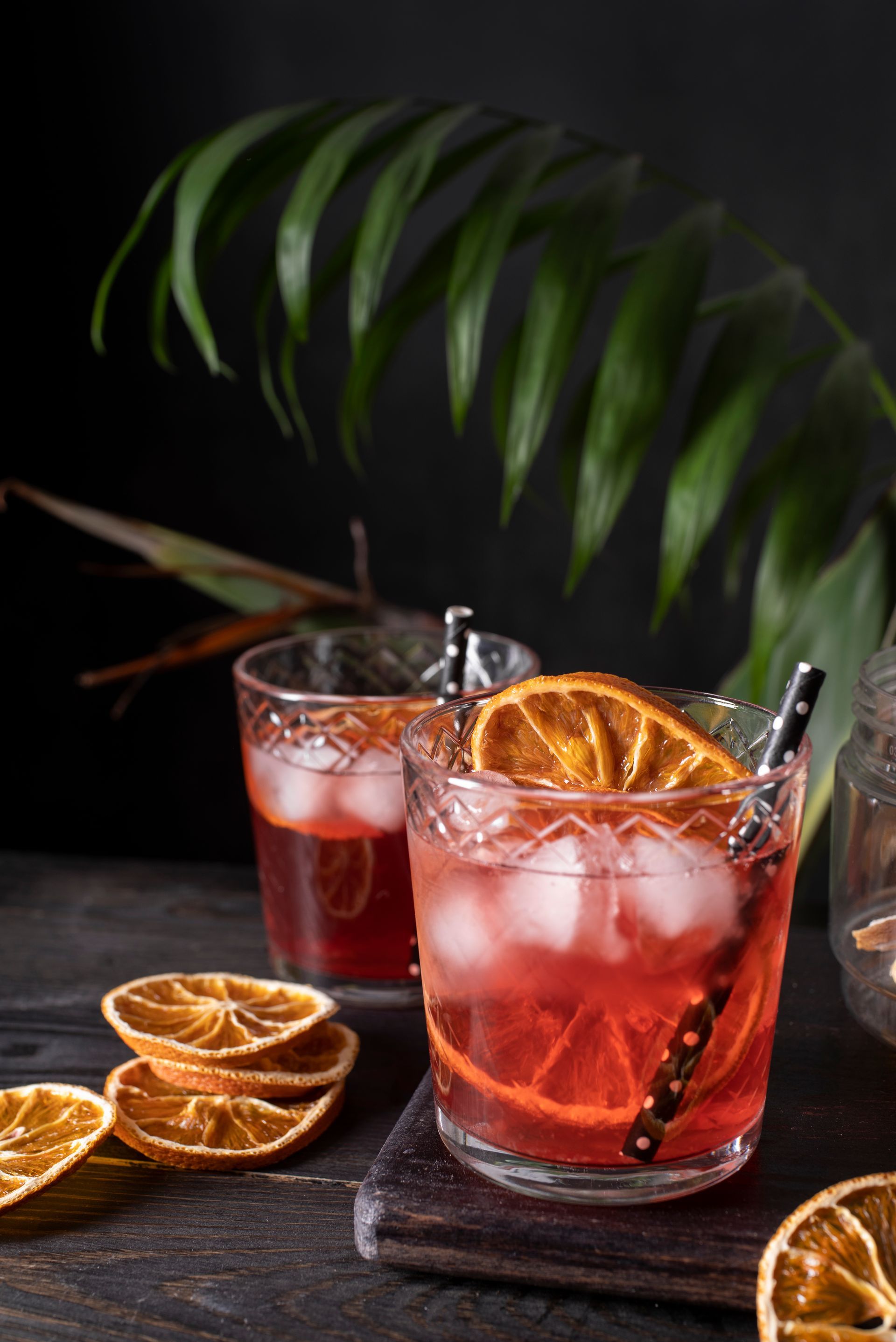 Due cocktail con ghiaccio e fette d'arancia, su uno sfondo scuro con una foglia di palma.