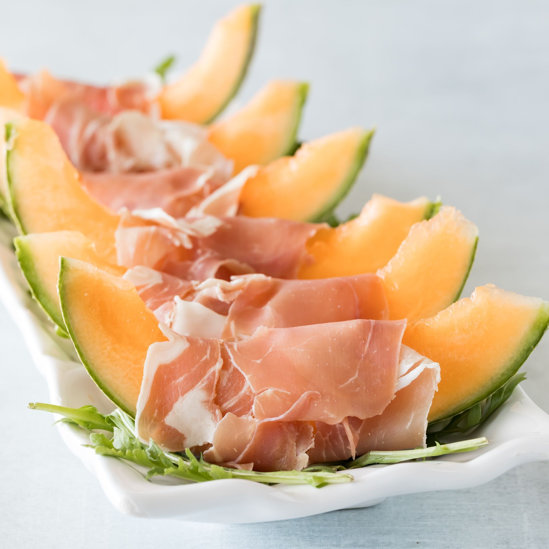 Fette di melone cantalupo con prosciutto su un piatto bianco rettangolare con rucola.