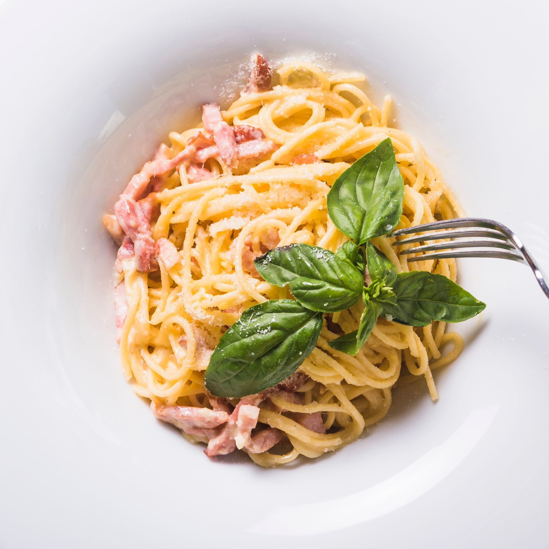 Pasta alla carbonara in una ciotola bianca, guarnita con basilico e parmigiano, condita con una forchetta.