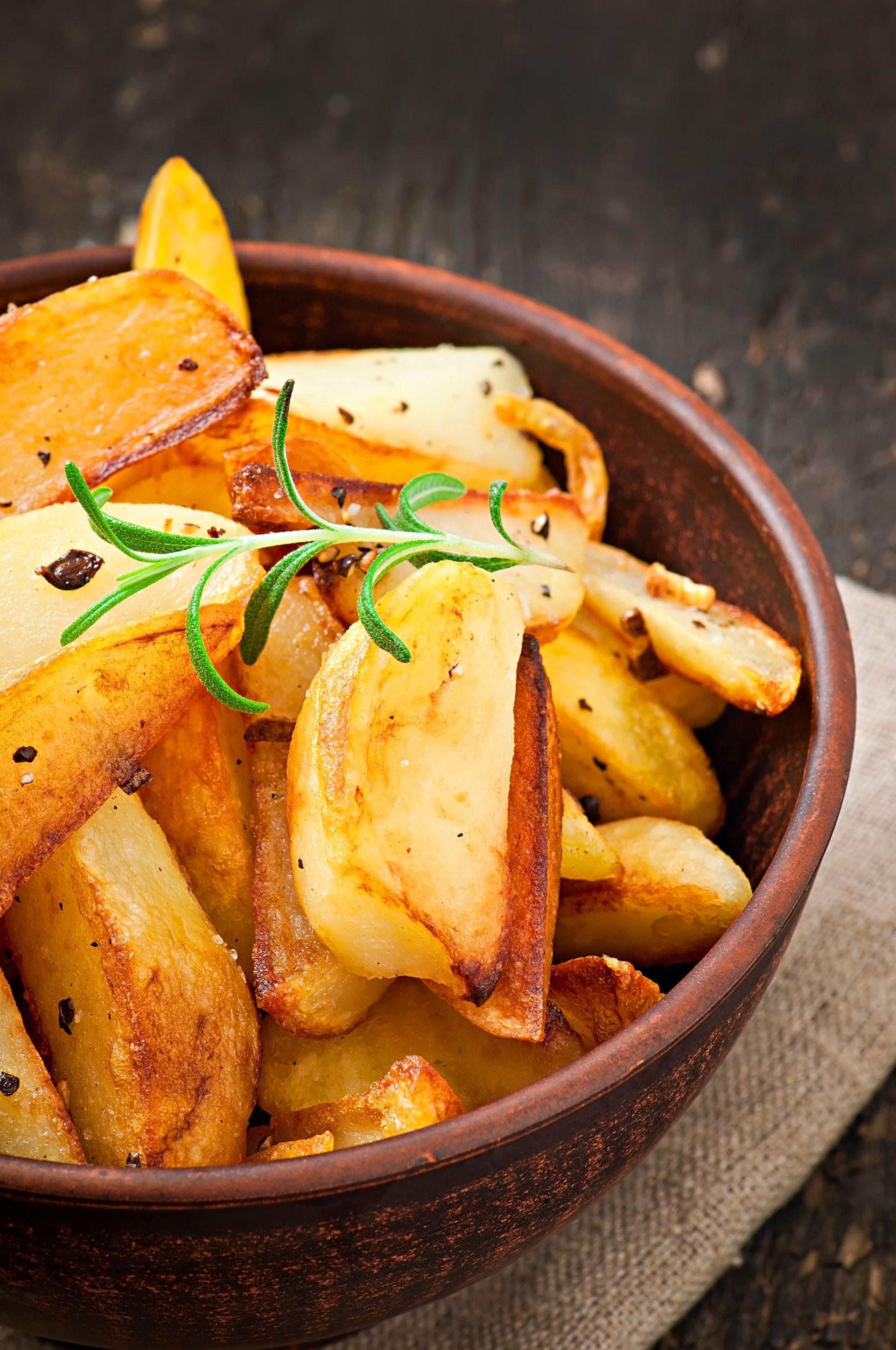 Spicchi di patate arrostite in una ciotola marrone, guarnite con rosmarino, su una superficie di legno rustica.