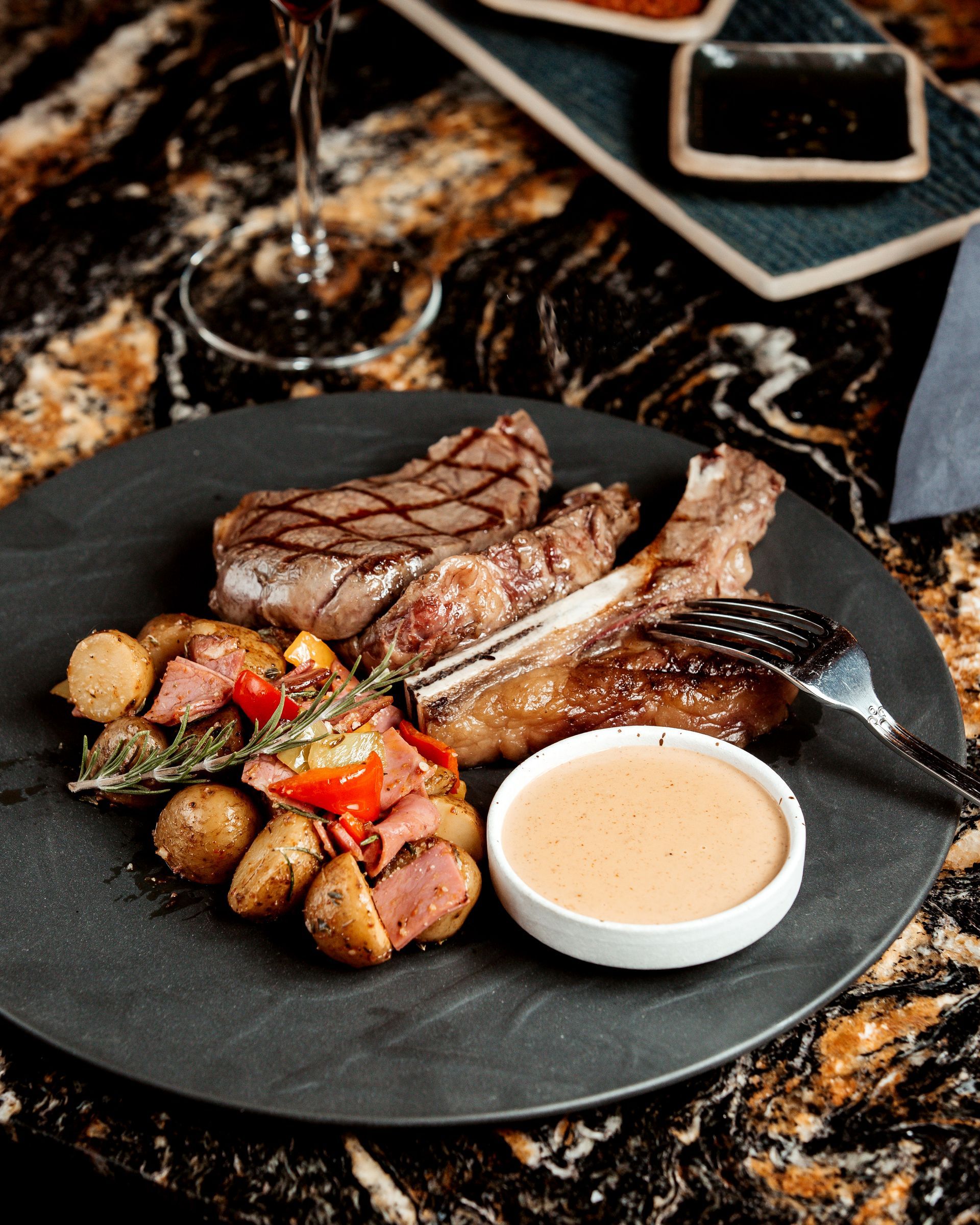 Bistecca e patate con salsa su un piatto nero, un bicchiere di vino e spezie su un piano di lavoro decorato.