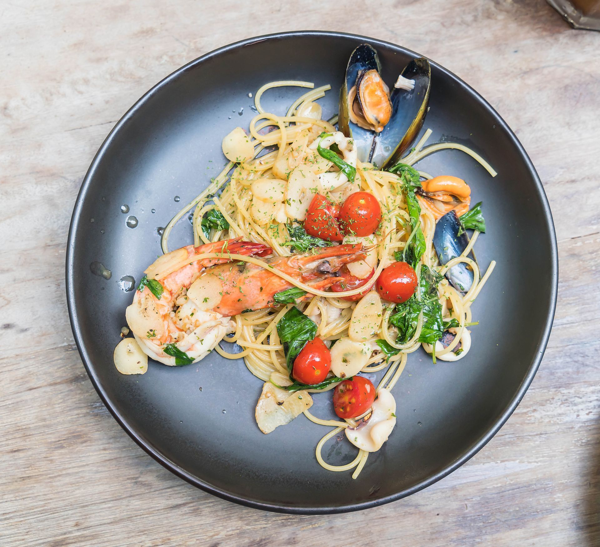 Pasta con gamberi, cozze, pomodorini ed erbe aromatiche su un piatto nero.
