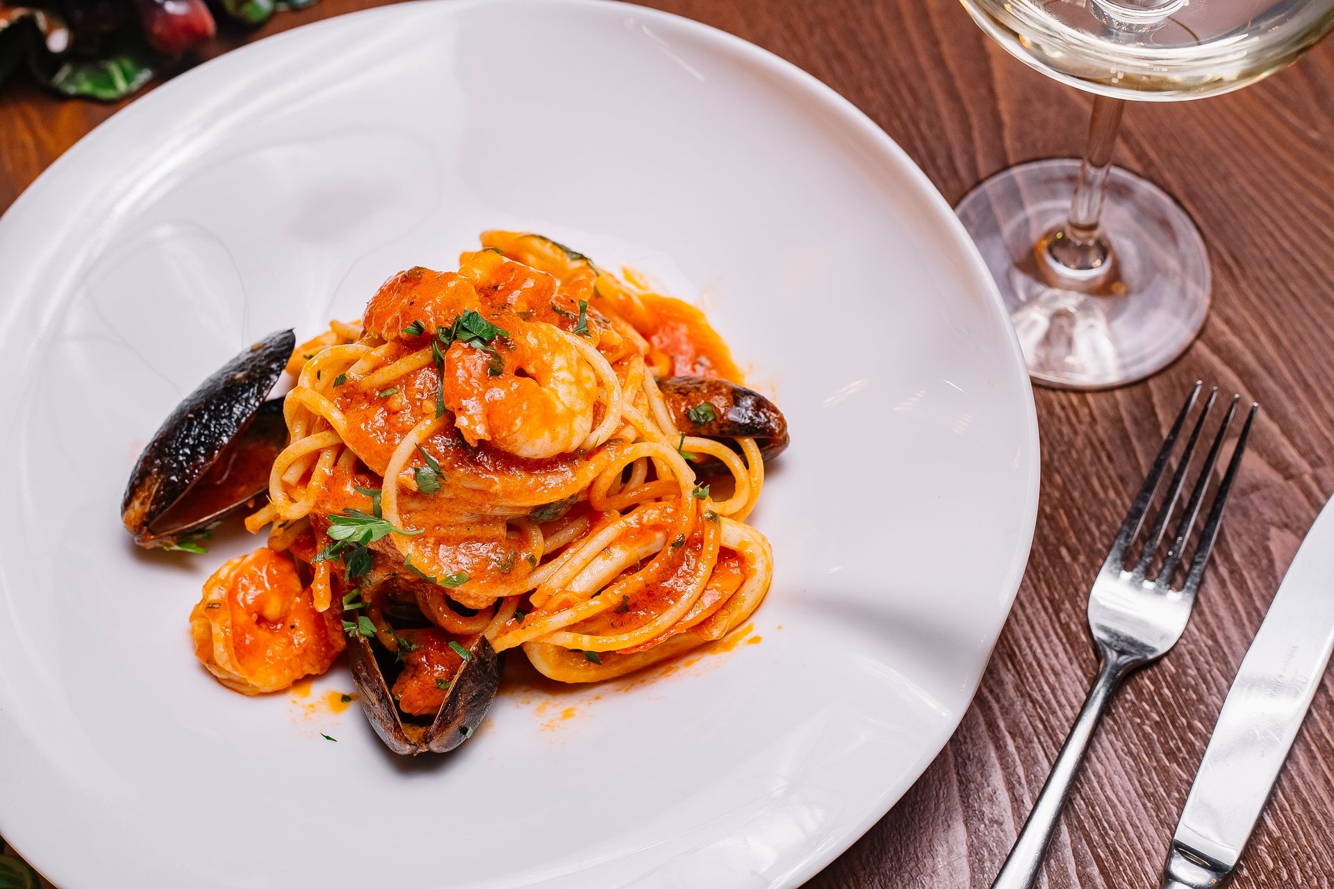 Pasta ai frutti di mare su un piatto bianco, con bicchiere di vino, forchetta e coltello su un tavolo di legno.