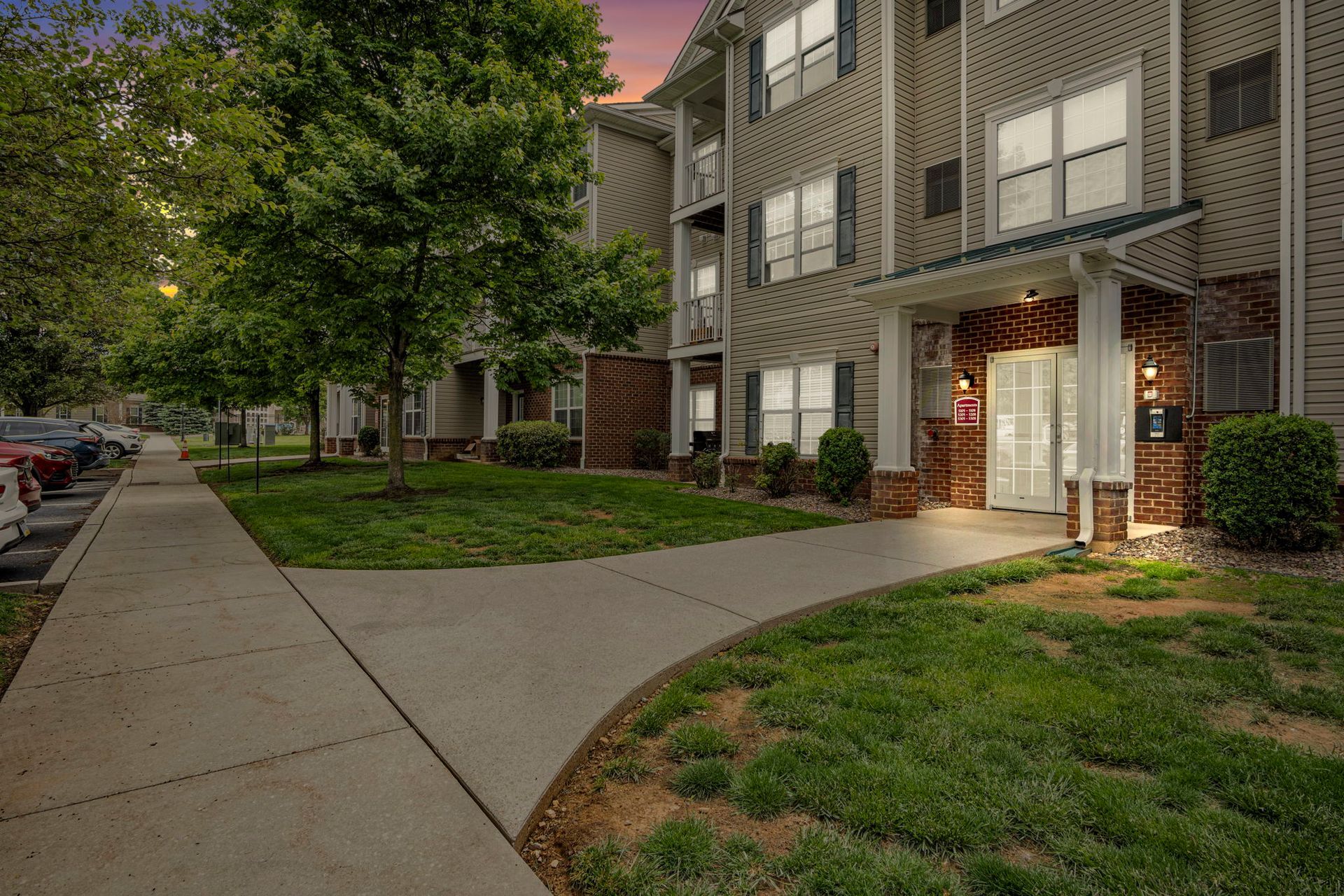 Apartment building entrance at dusk with lit doorway, trees, sidewalk, and grassy lawn