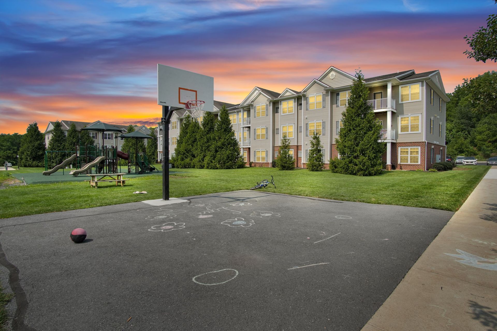 Outdoor basketball court beside apartment buildings at sunset, with a basketball on the pavement.