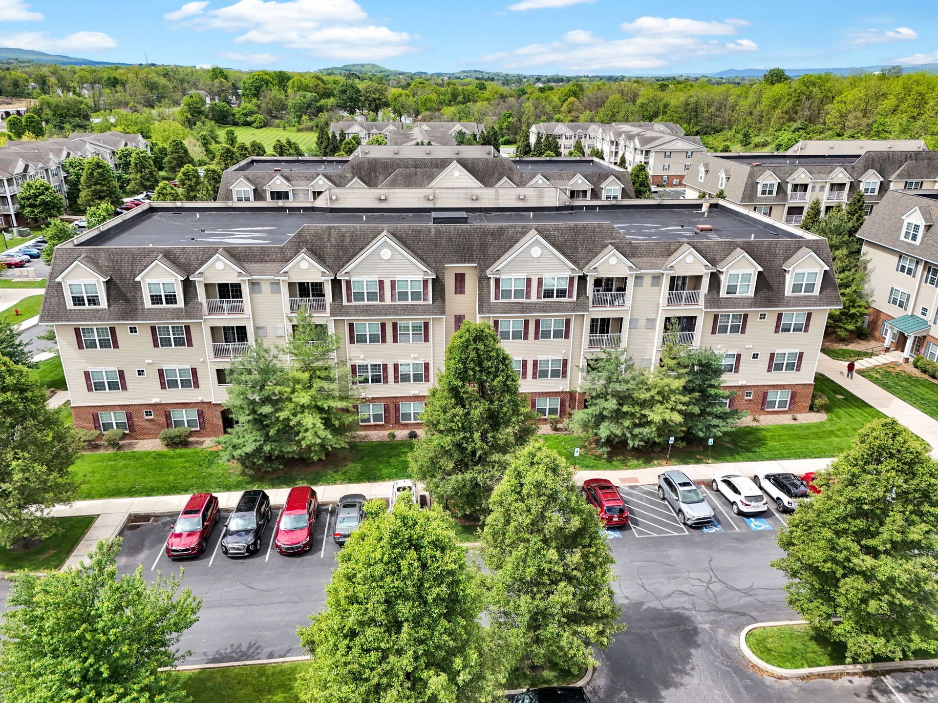 Aerial view of a large apartment building with parked cars and trees on a sunny day