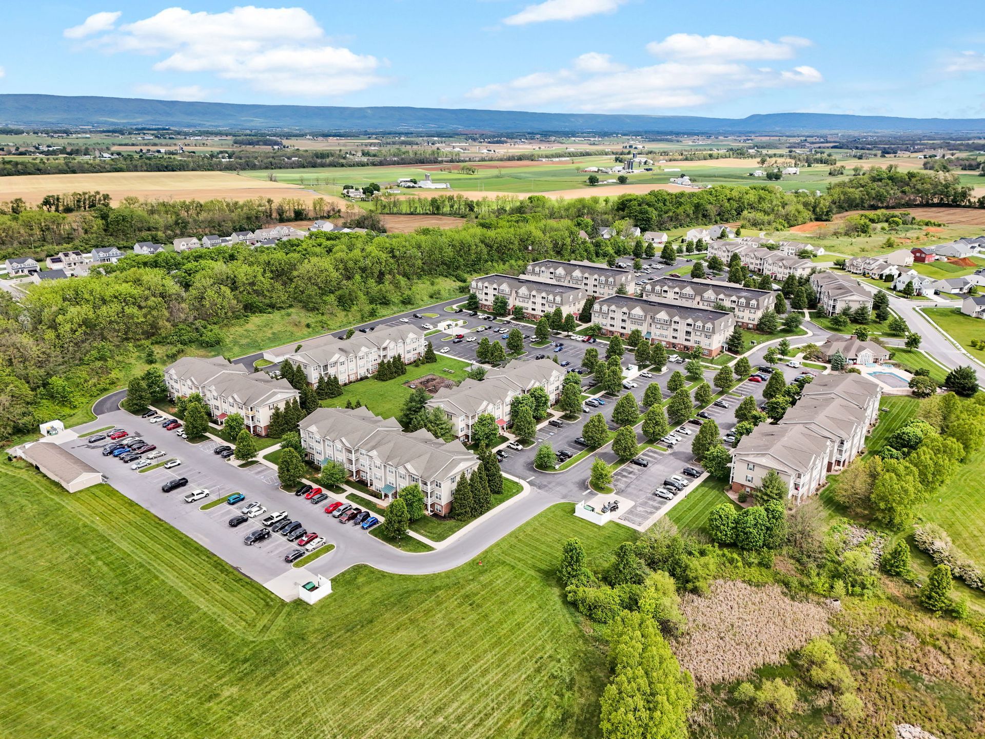 Aerial view of a suburban apartment complex surrounded by green fields and trees under a bright blue sky