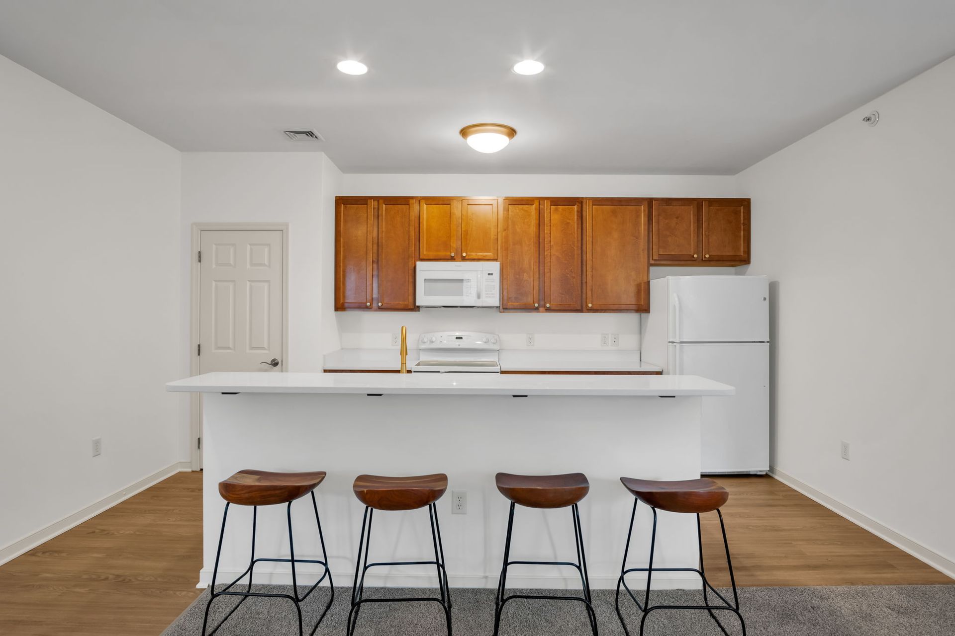 Empty kitchen with white island, wooden cabinets, and four stools in a bright open room