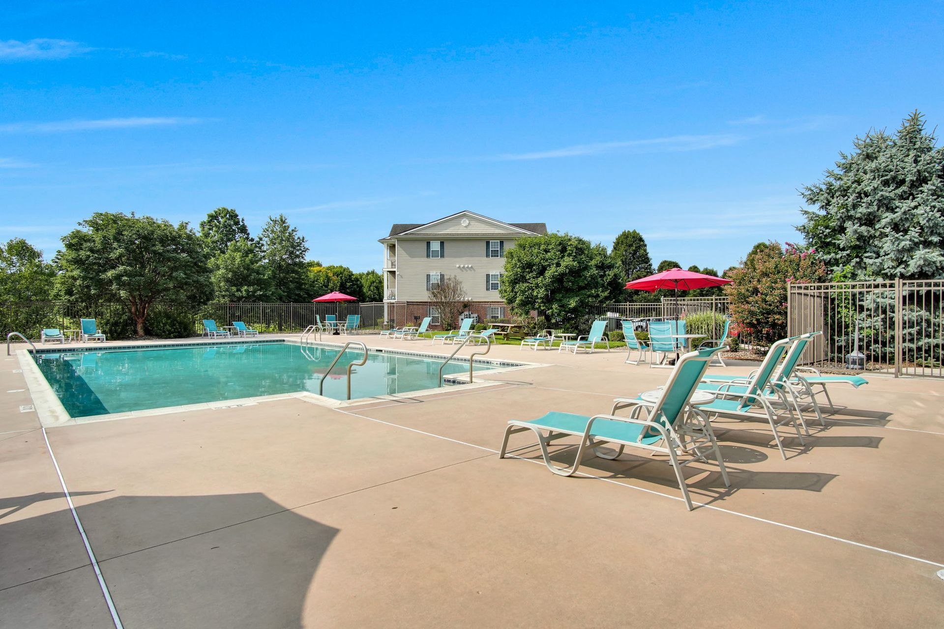 Outdoor swimming pool with lounge chairs, red umbrellas, and a clubhouse under a blue sky