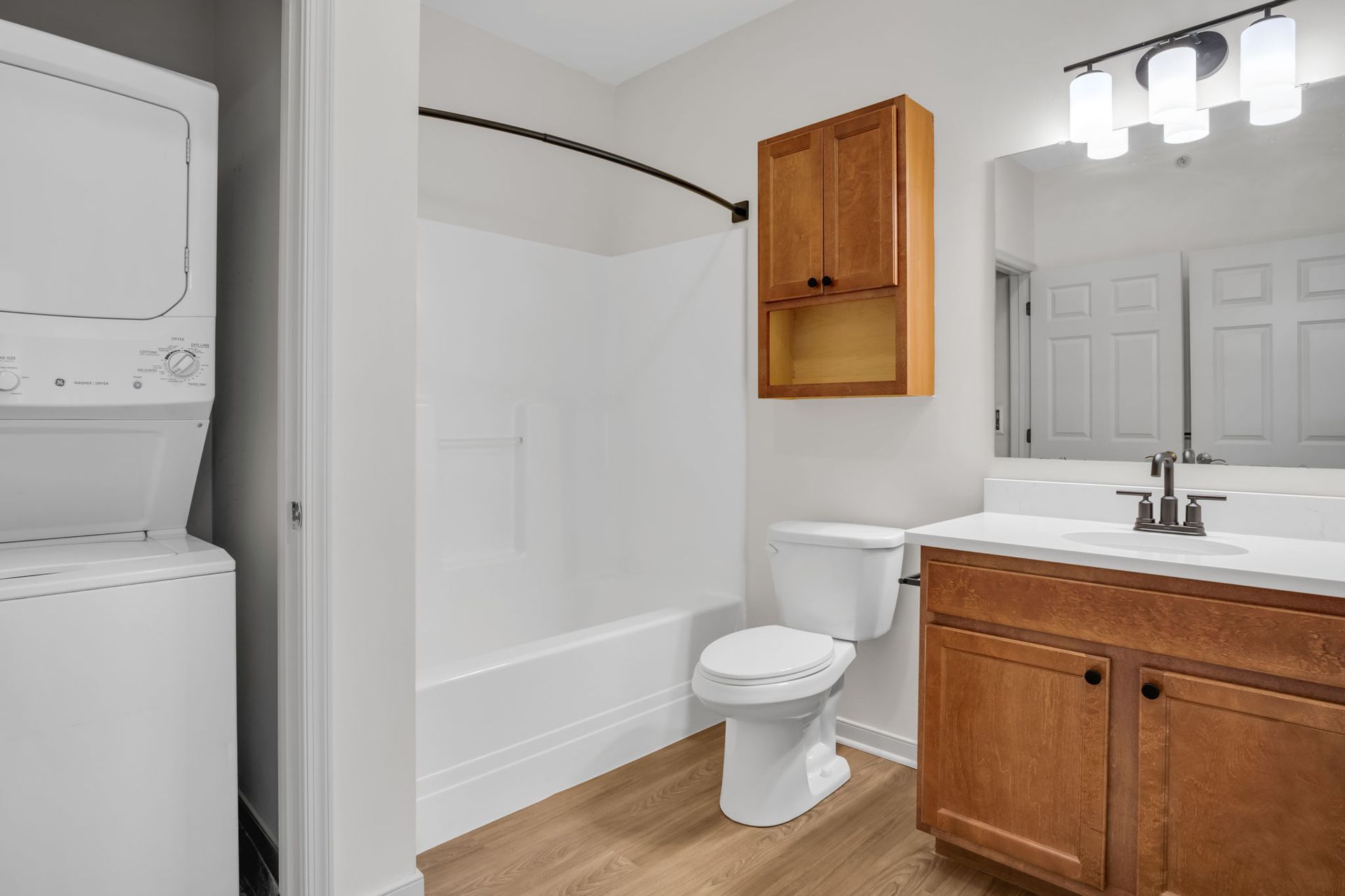 Small bathroom with stacked washer/dryer, tub, toilet, wood vanity, and mirror.