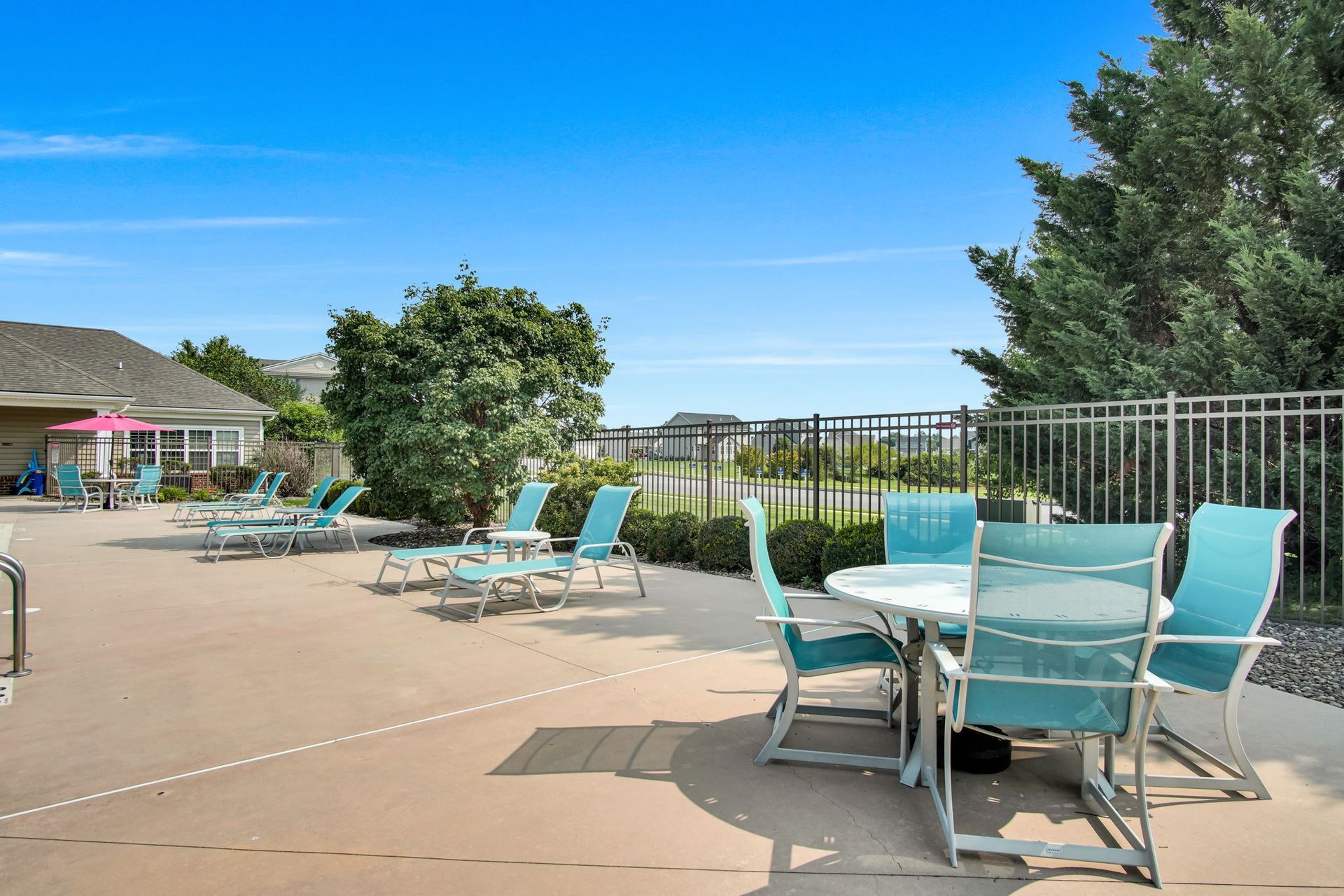 Pool deck with turquoise lounge chairs, tables, and umbrellas beside a fenced water view under a bright blue sky