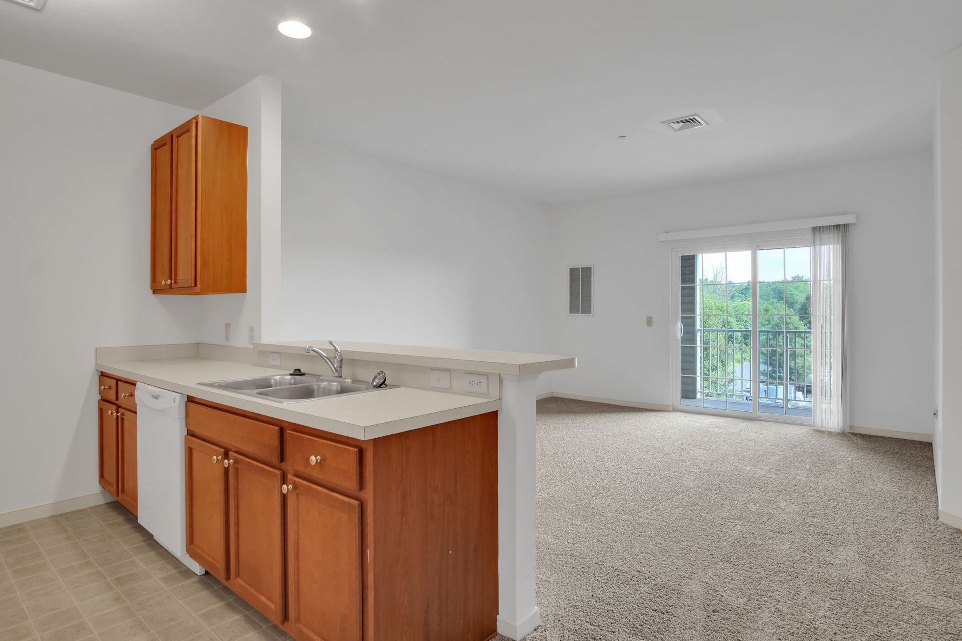 Empty apartment kitchen with wood cabinets and carpeted living area leading to sliding glass doors