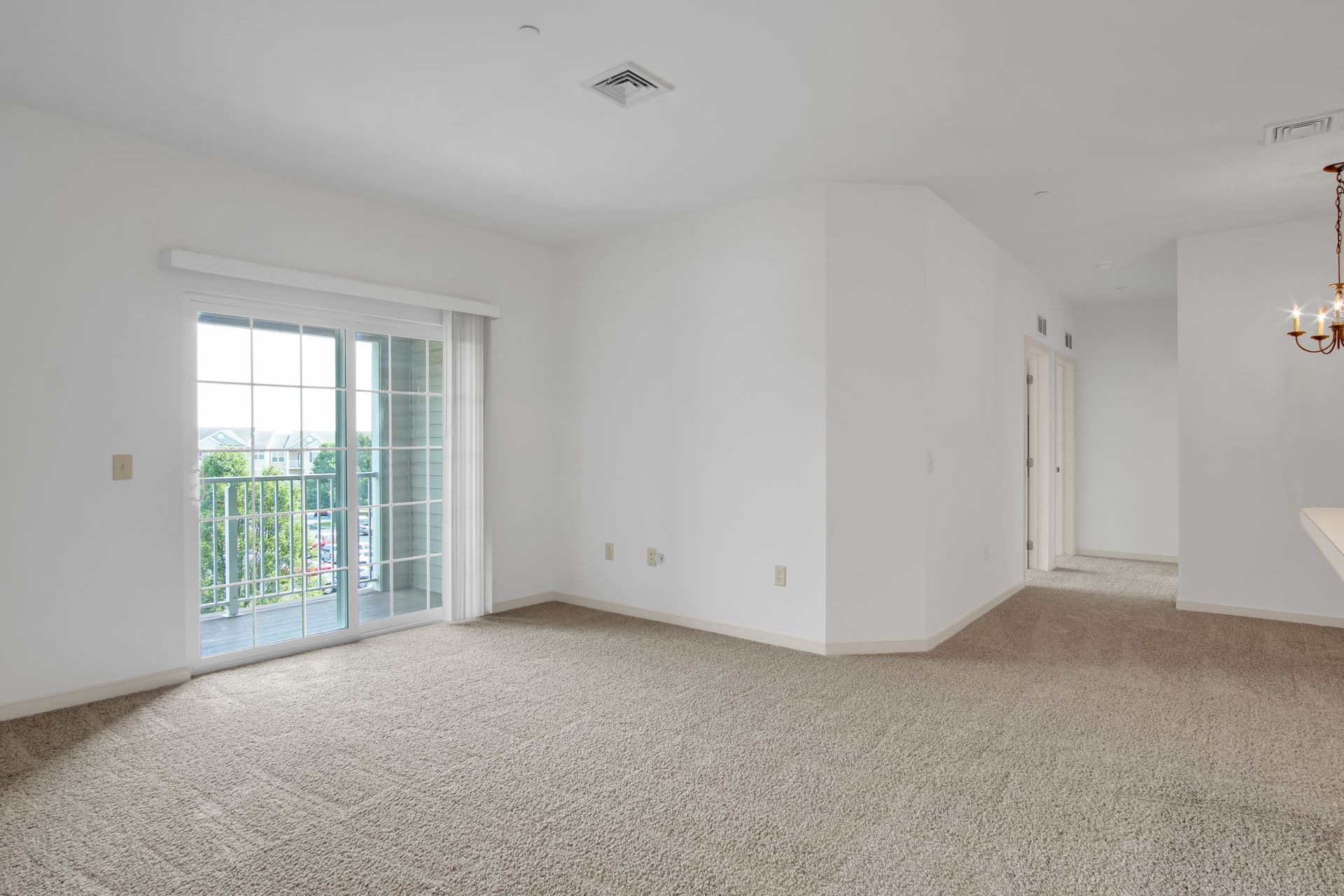 Empty carpeted living room with white walls, sliding glass doors, and an open hallway