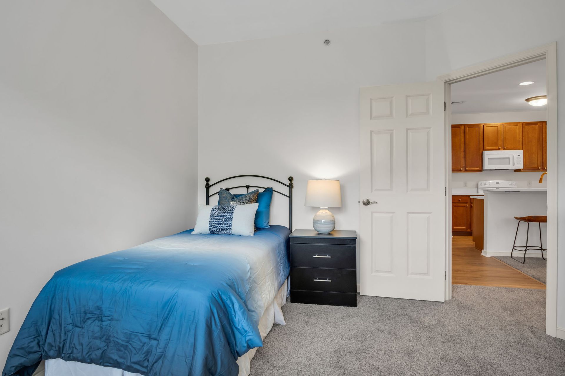 Small bedroom with a blue bedspread, black nightstand, and open door to a kitchen.