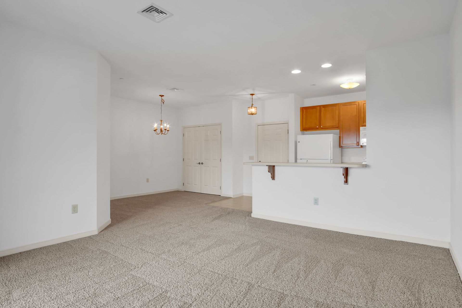 Empty beige carpeted living room with white walls and a small white kitchen pass-through.